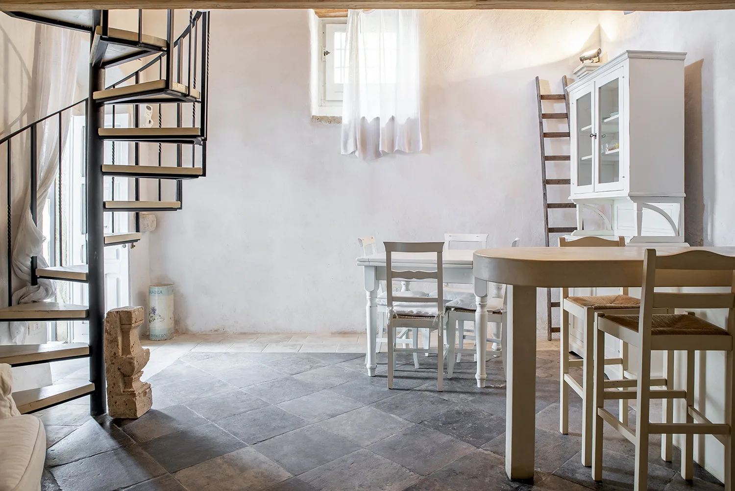 A bright dining area with a table, chairs, and a narrow spiral staircase.