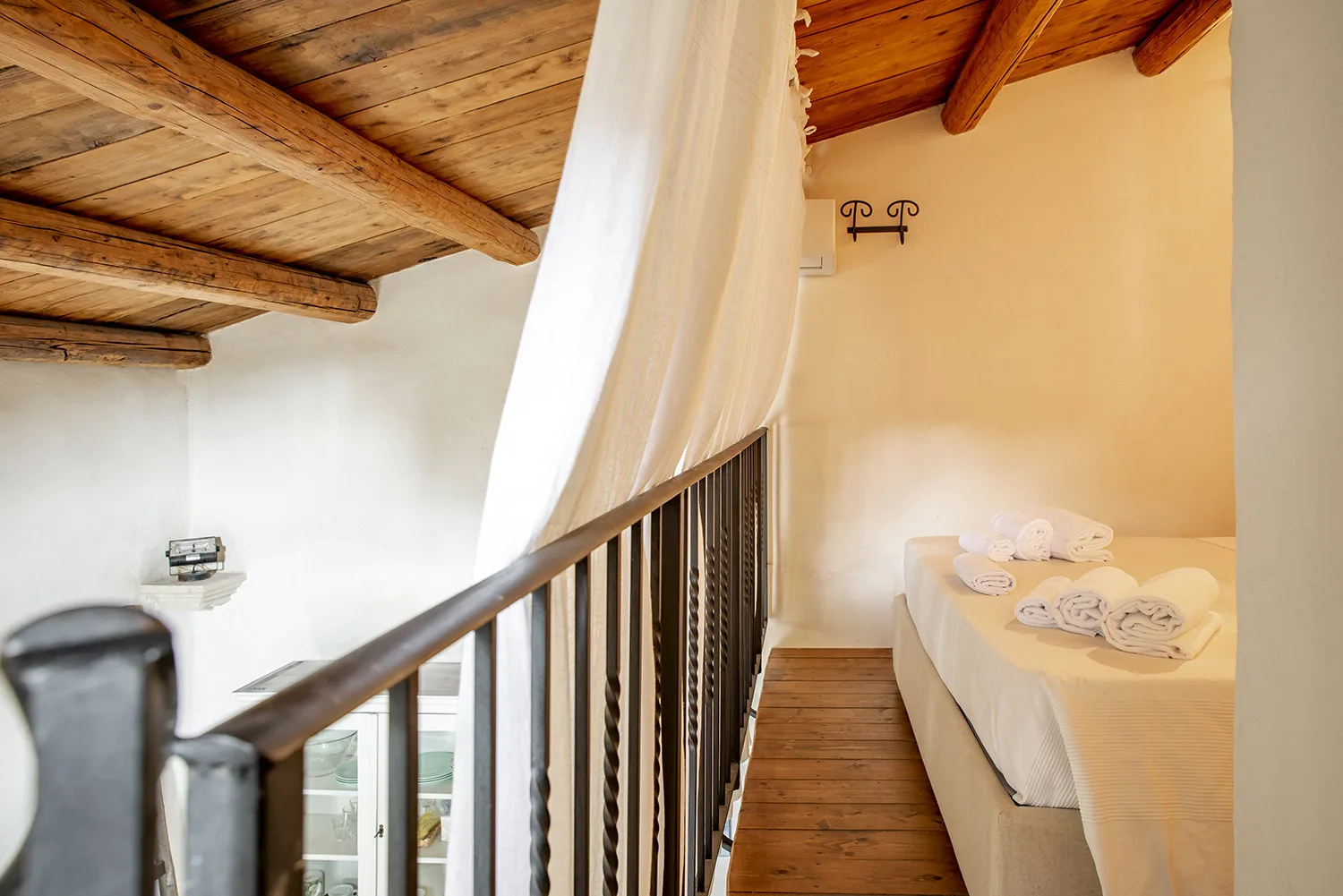 A loft bedroom with a bed beside a wooden railing and exposed ceiling beams.