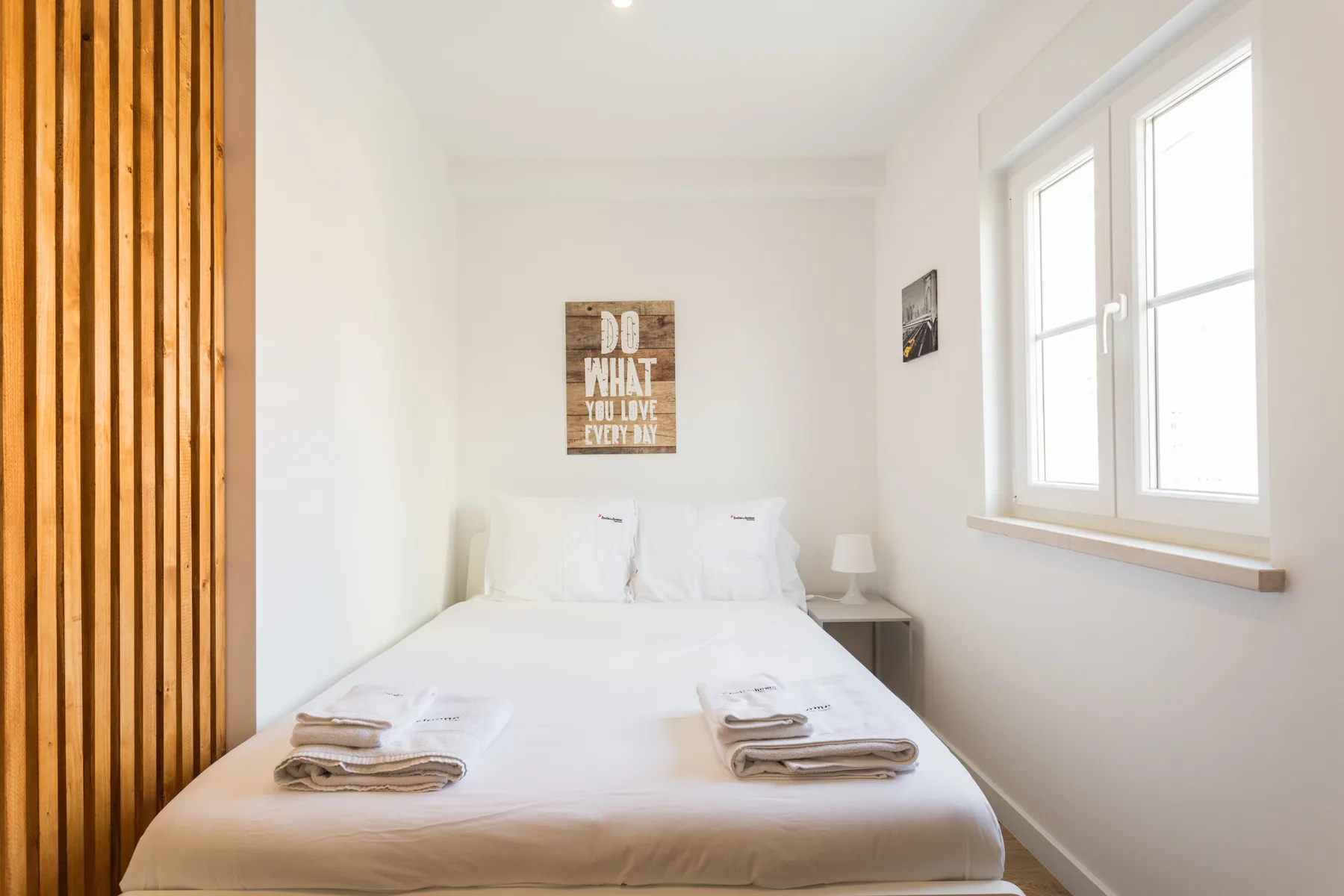 A bright bedroom with white bedding and a large window.