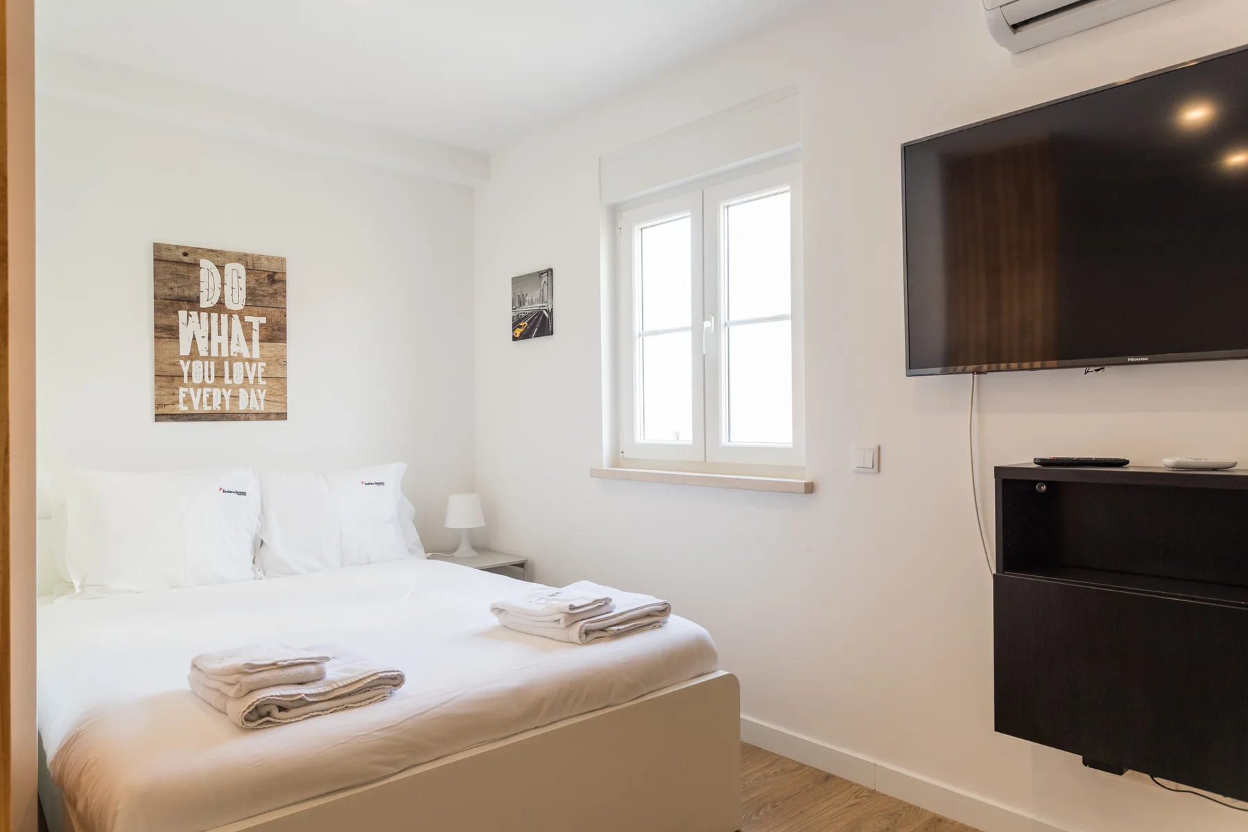A bright bedroom with a made bed, window, and wall-mounted television.