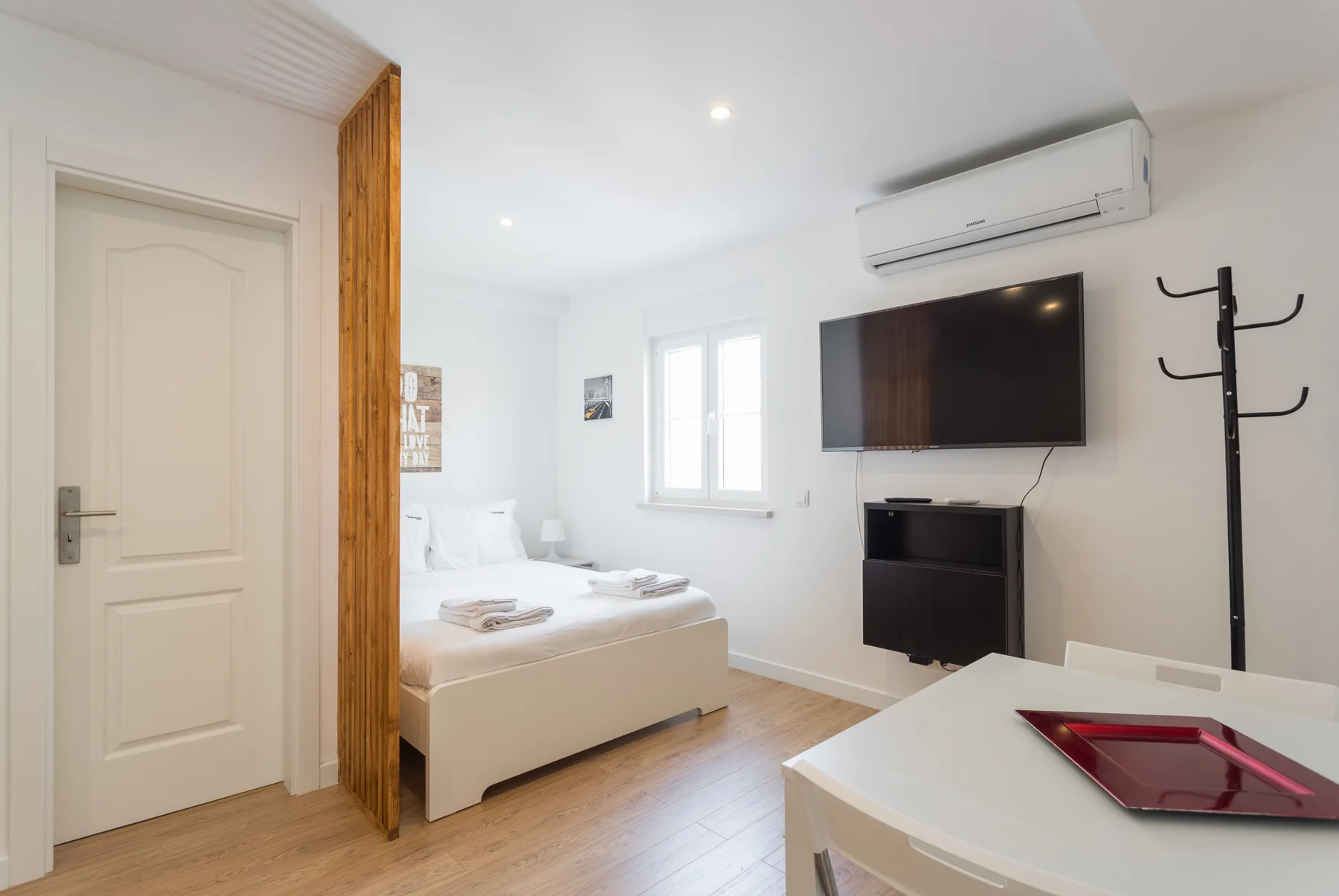 Un estudio luminoso con una cama, un televisor en la pared y una mesa de comedor.