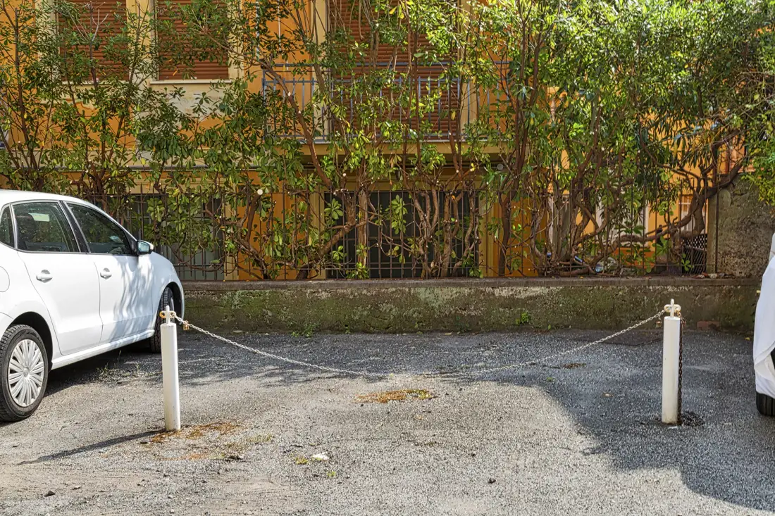 Un área de estacionamiento de grava está frente a la fachada frondosa de un edificio.