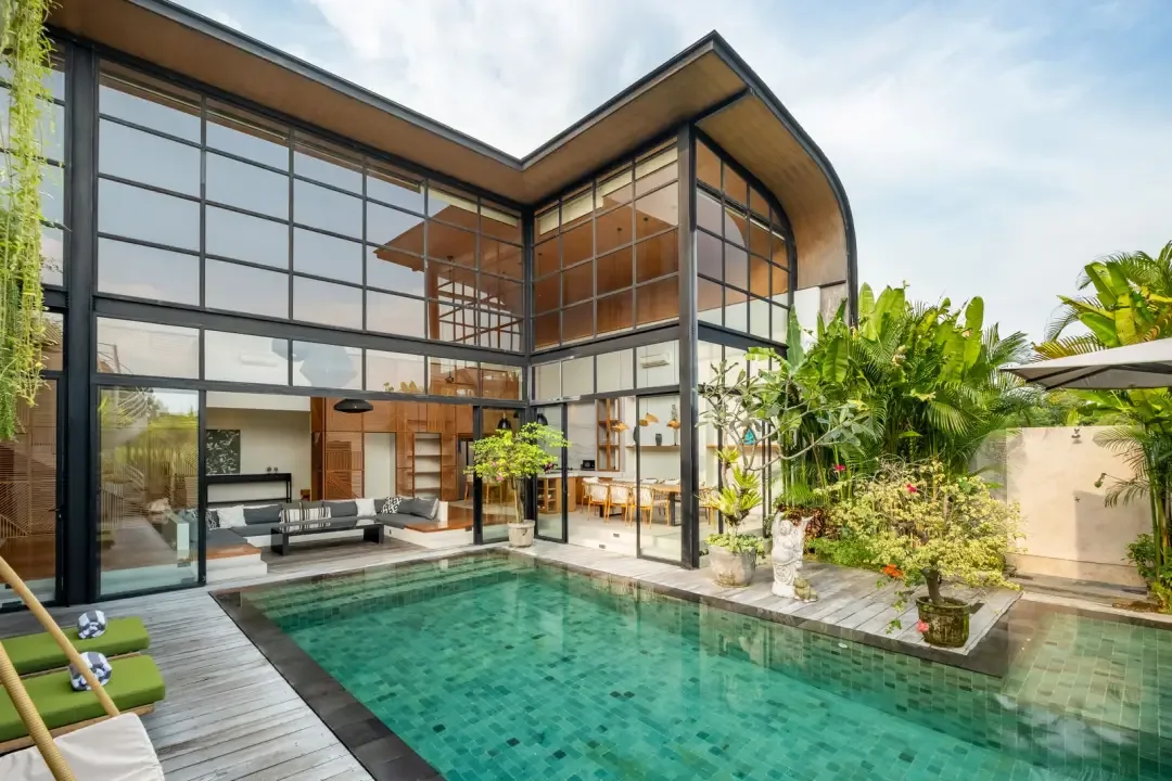 A modern glass-fronted villa overlooks a swimming pool and tropical garden.