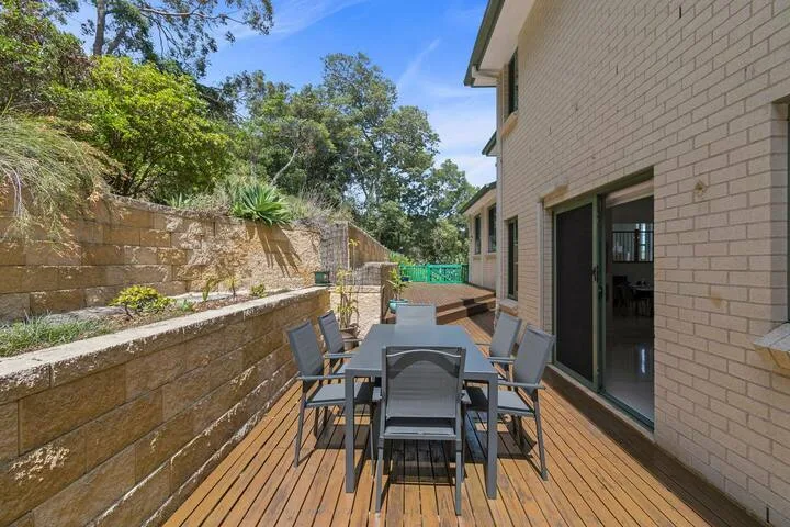 Una zona de comedor al aire libre con mesa y sillas en una terraza de madera junto a un edificio de ladrillo.