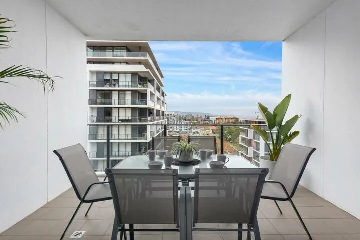 Un balcón cubierto tiene una mesa de comedor y asientos con vista a edificios de apartamentos cercanos.