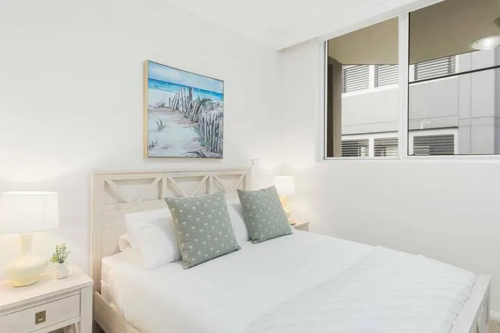 Un dormitorio luminoso con una cama bien tendida y una lámina enmarcada de una playa.