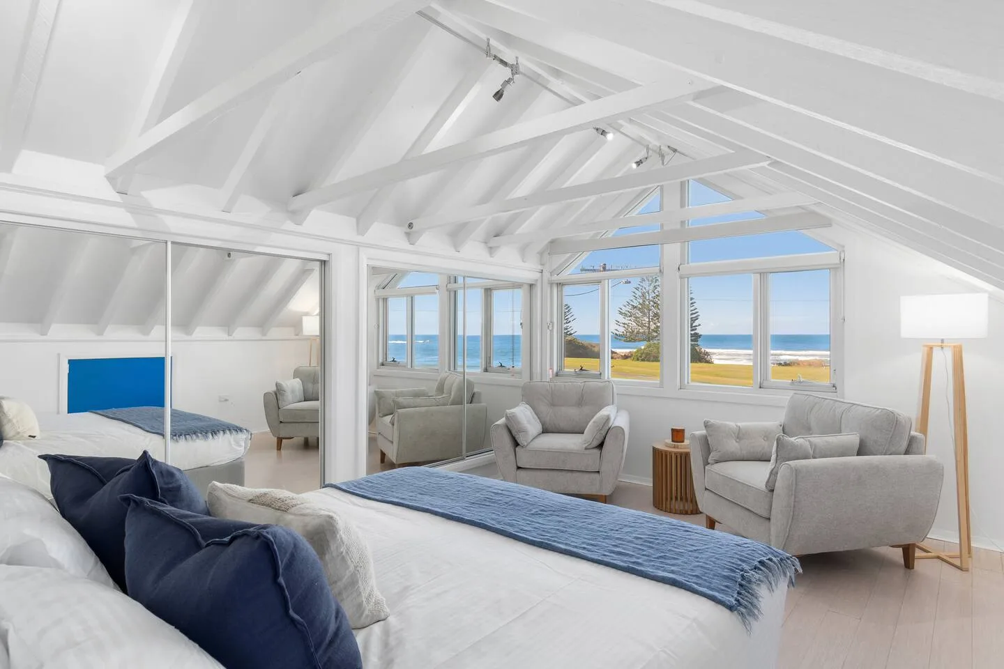 Un dormitorio luminoso con cama y asientos junto a grandes ventanas con vistas al océano.