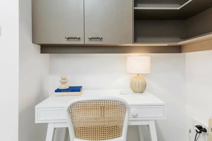 Un pequeño escritorio está bajo armarios de pared, con una silla tejida y una lámpara de mesa.