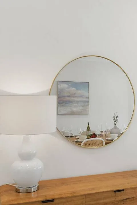 Cómoda de dormitorio con espejo redondo, lámpara y un paisaje marino enmarcado.
