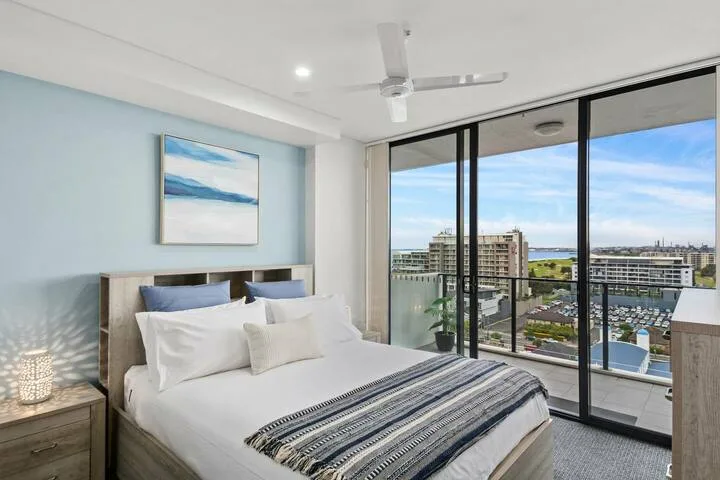 Un dormitorio con una cama bien tendida junto a grandes ventanales con vistas a la ciudad.