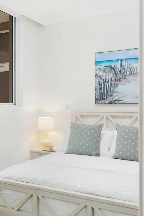 Un dormitorio luminoso con una cama blanca, cojines estampados y arte costero en la pared.