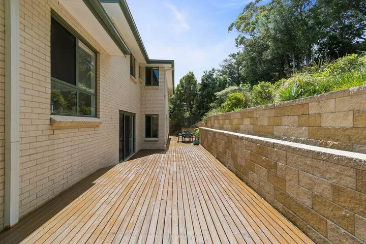 Una larga terraza de madera bordea un edificio de ladrillo claro y un muro de contención de piedra.