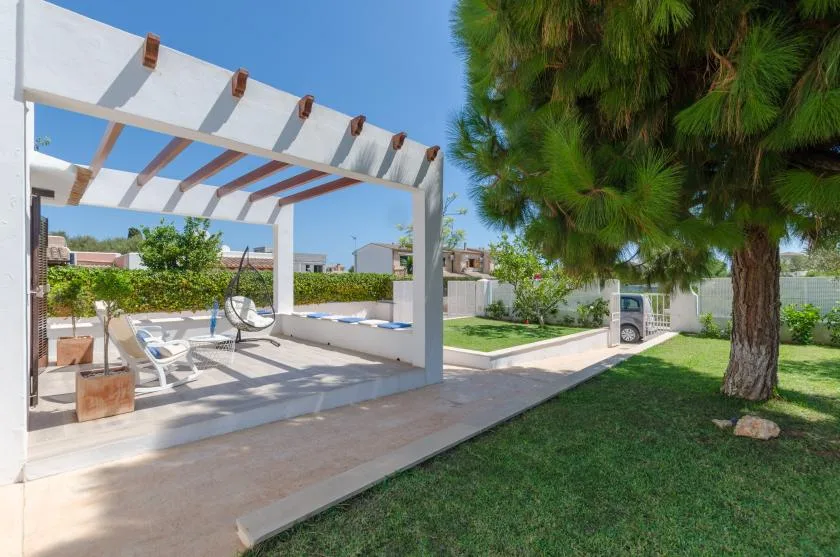 Un patio exterior con una pérgola blanca, asientos y un árbol junto al césped.