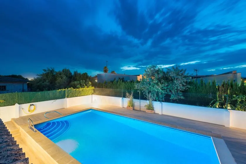Una piscina exterior iluminada está junto a una terraza pavimentada bajo un cielo al anochecer.