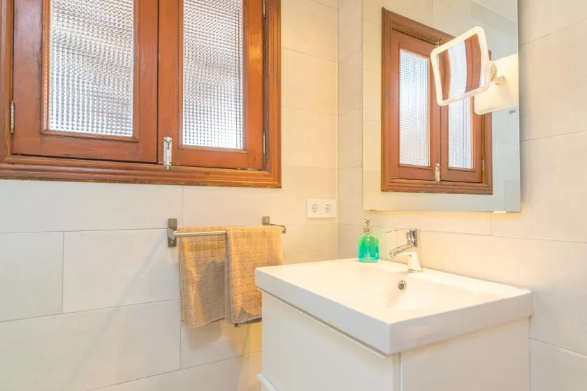 Un lavabo de baño está bajo un espejo, junto a una ventana con marco de madera.