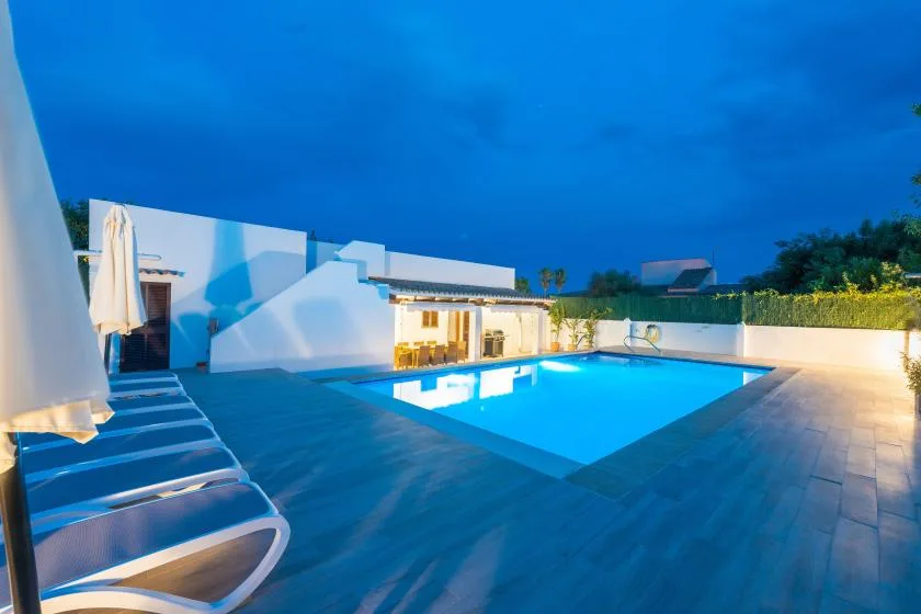 Una piscina al aire libre brilla junto a un patio pavimentado al anochecer.