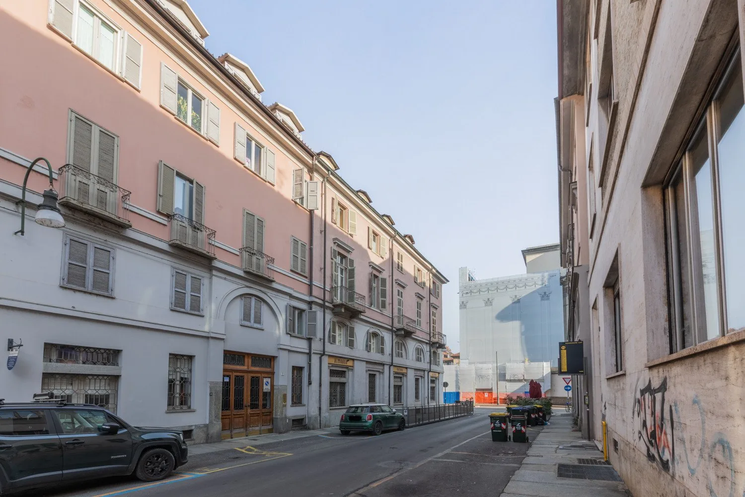 Una calle urbana pasa entre edificios de varias plantas, con coches estacionados junto a la acera.