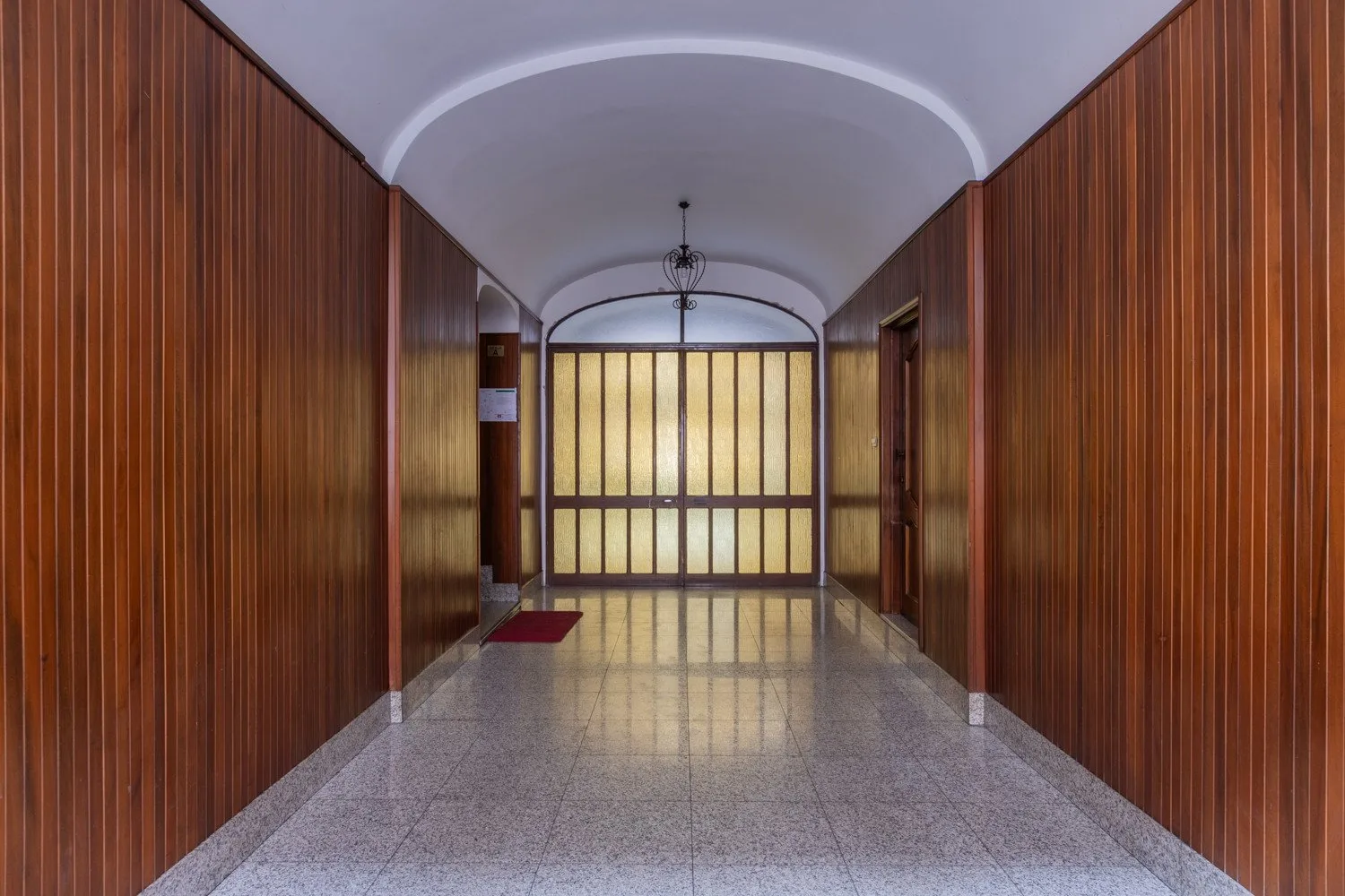 Un largo pasillo interior tiene paredes revestidas de madera y una puerta acristalada iluminada al fondo.