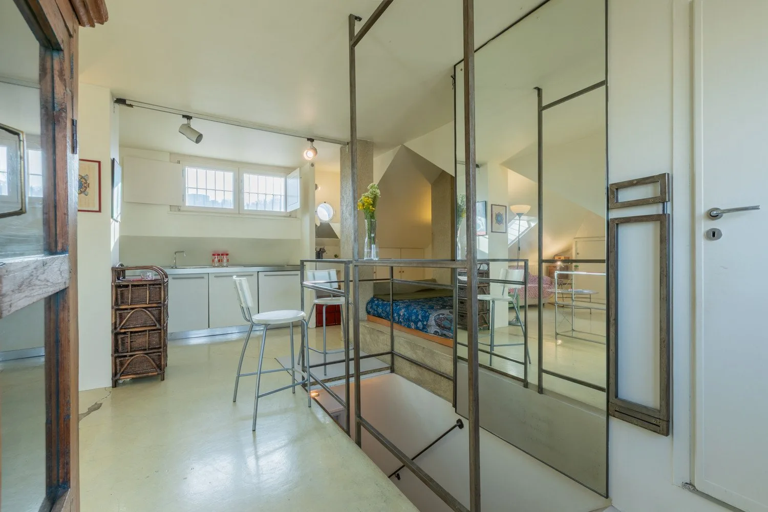Un estudio luminoso con una pequeña cocina, una mesa y una cama tras una mampara de vidrio.