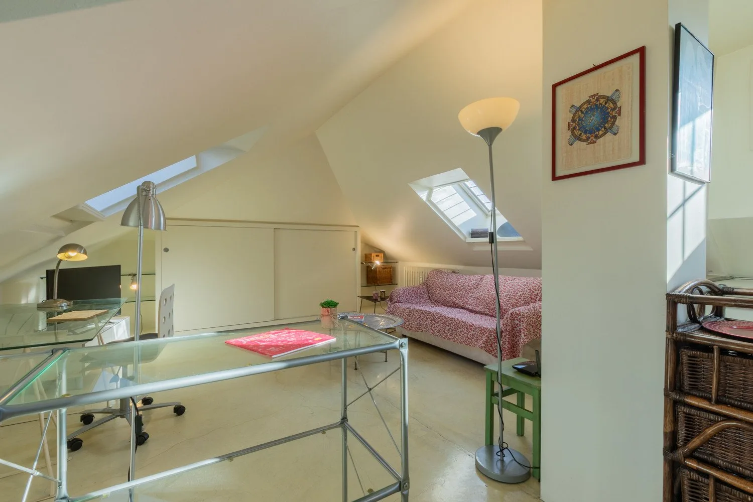 Una luminosa habitación abuhardillada con escritorio de vidrio, asientos y techo inclinado.