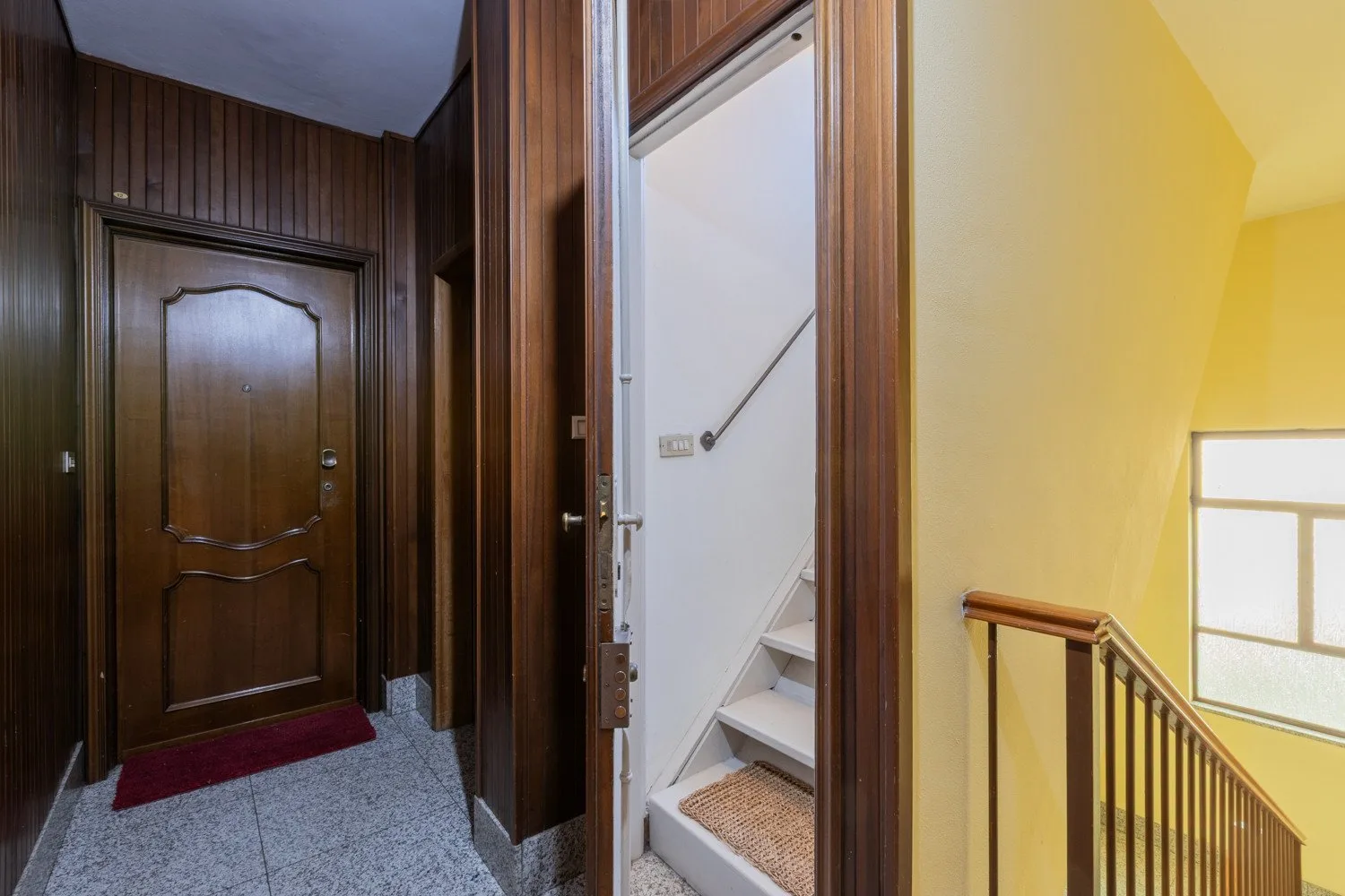 Una entrada revestida de madera da a una escalera junto a una pared amarilla iluminada.