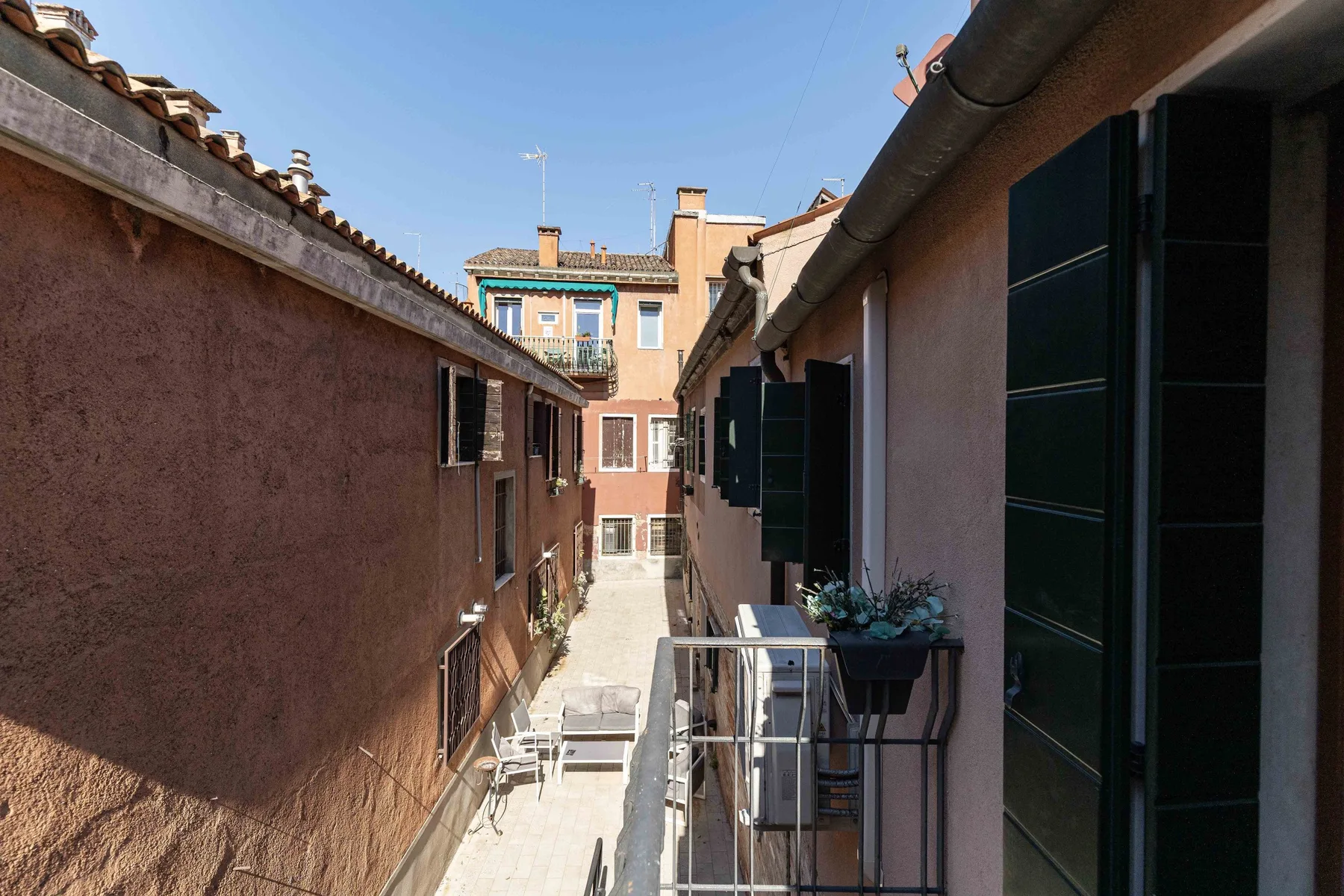 Un pasaje exterior estrecho entre edificios tiene contraventanas abiertas y una barandilla.