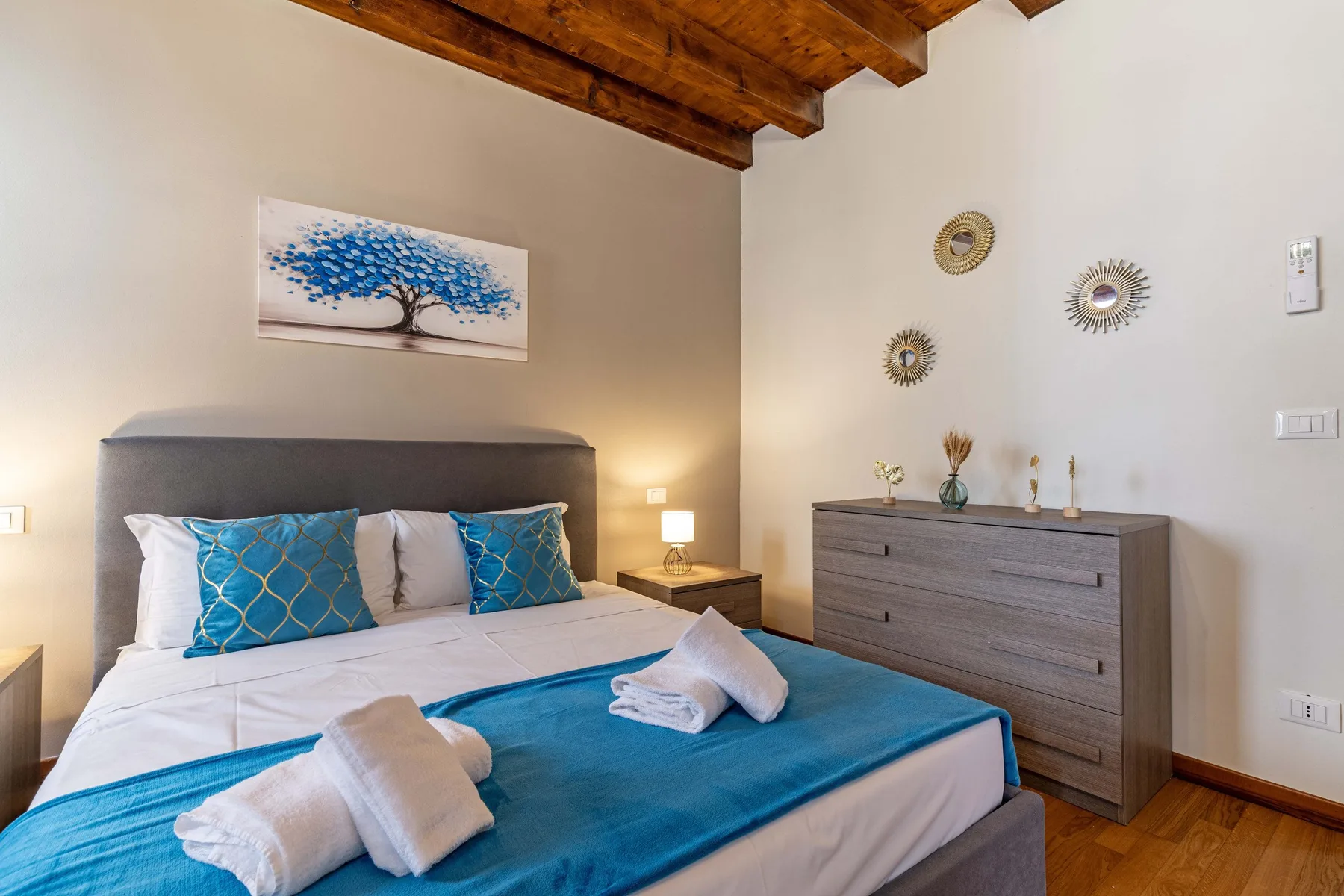 Dormitorio con una cama bien hecha y ropa de cama azul bajo vigas de madera vistas.