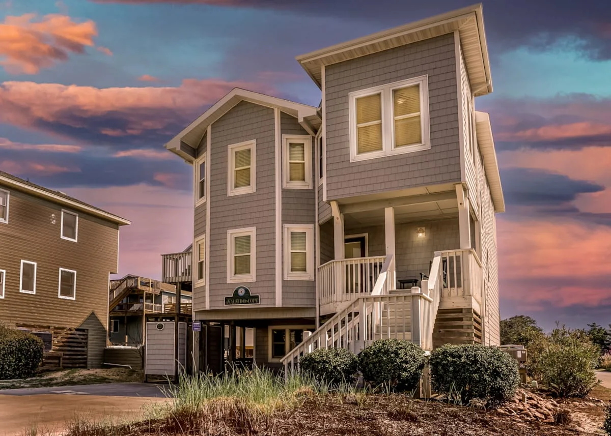 A raised gray house with exterior stairs stands beneath a colorful sunset sky.