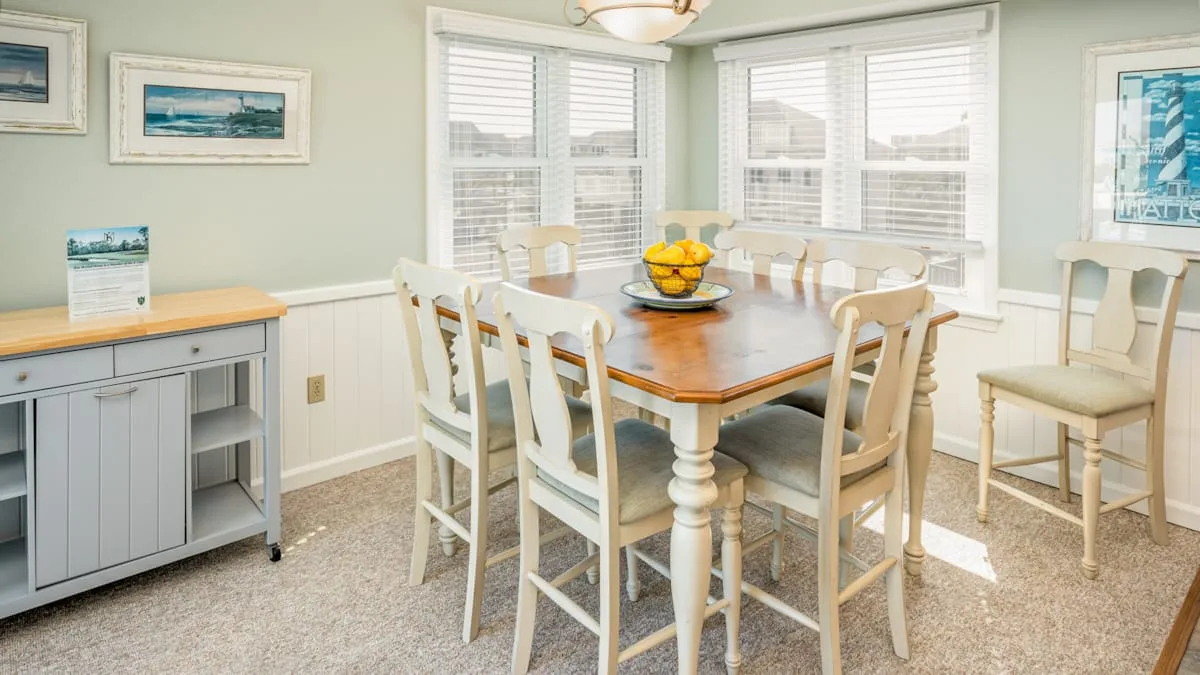 Un comedor luminoso con una mesa de madera, asientos blancos y ventanas grandes.