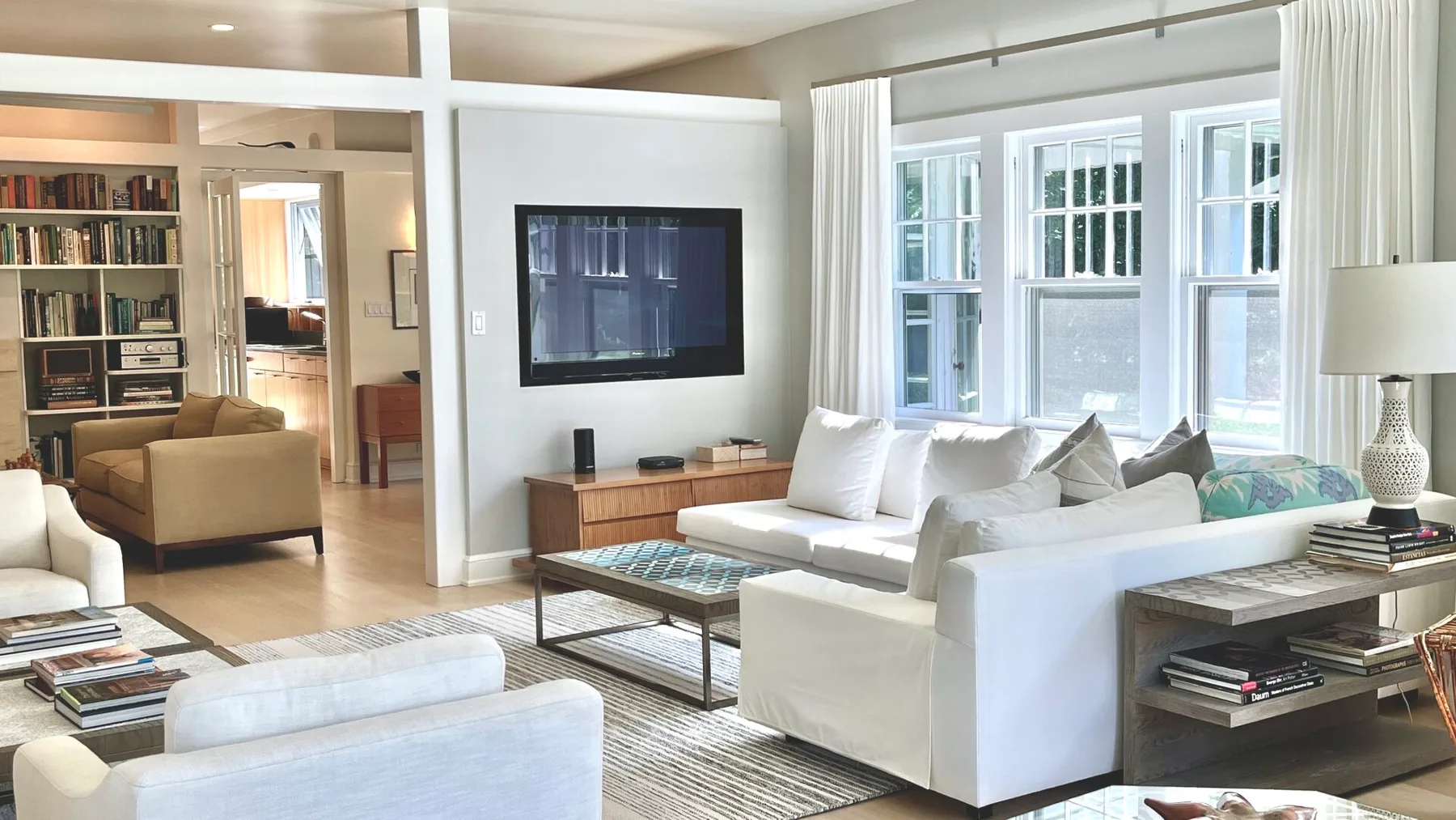 Una sala de estar luminosa con asientos blancos, ventanas grandes y un televisor en la pared.
