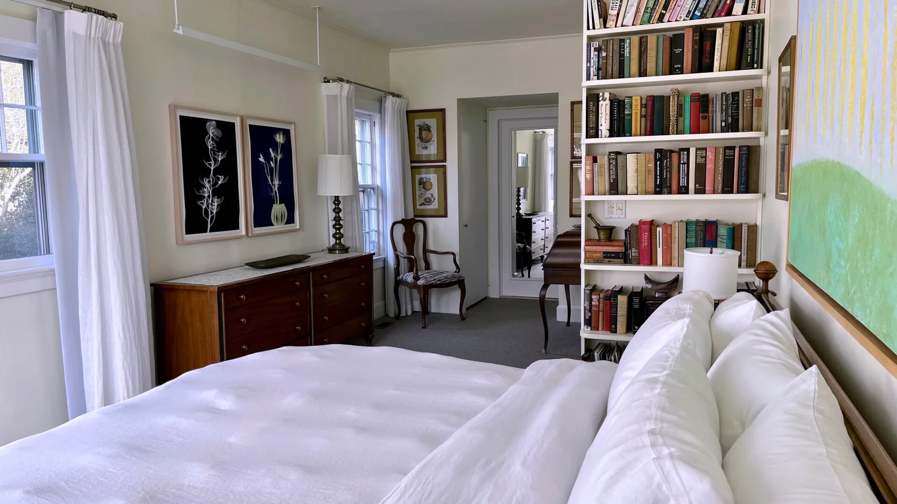 Un dormitorio luminoso con una cama blanca, una librería y ventanas.