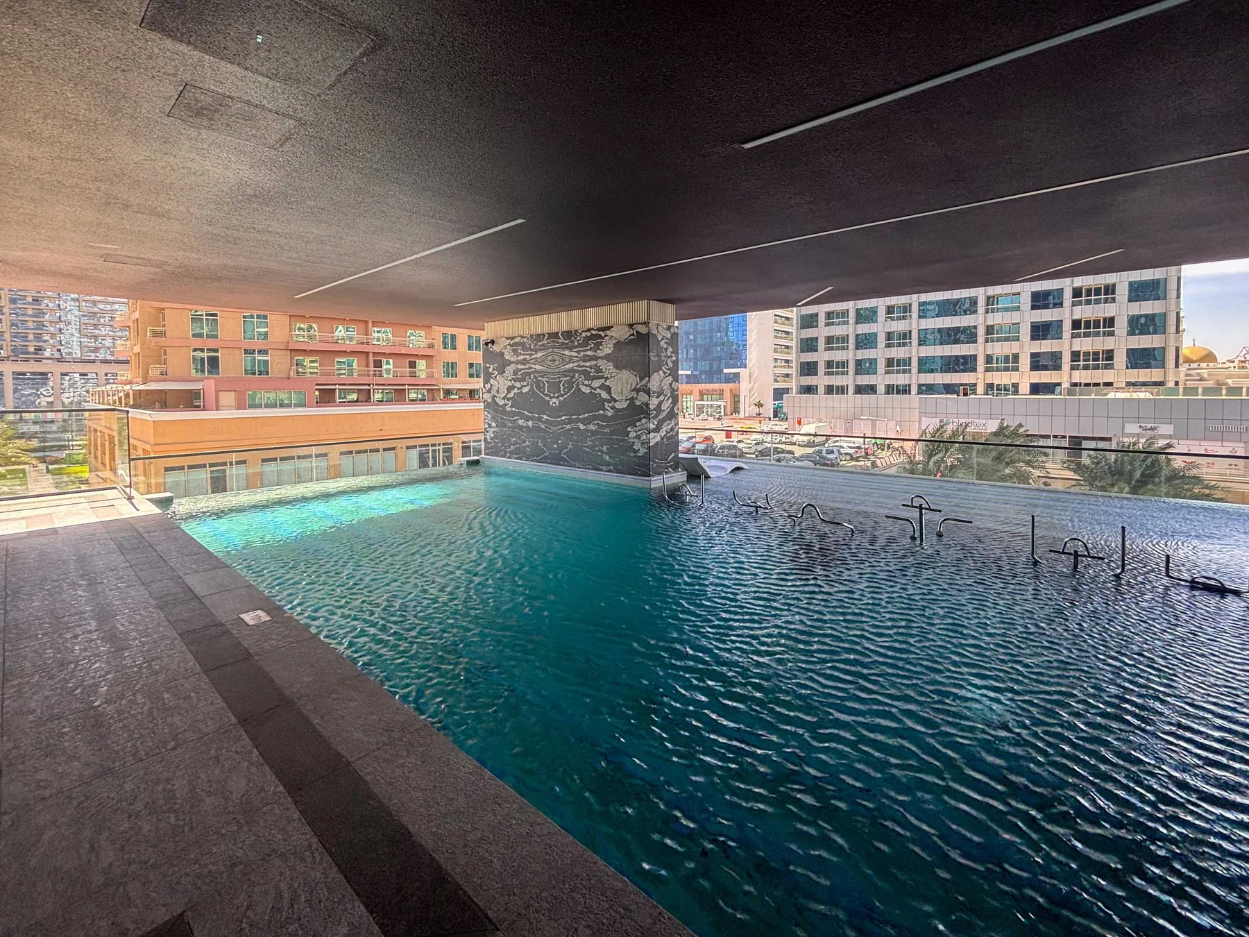 Una piscina está bajo una terraza cubierta, con edificios urbanos visibles al fondo.