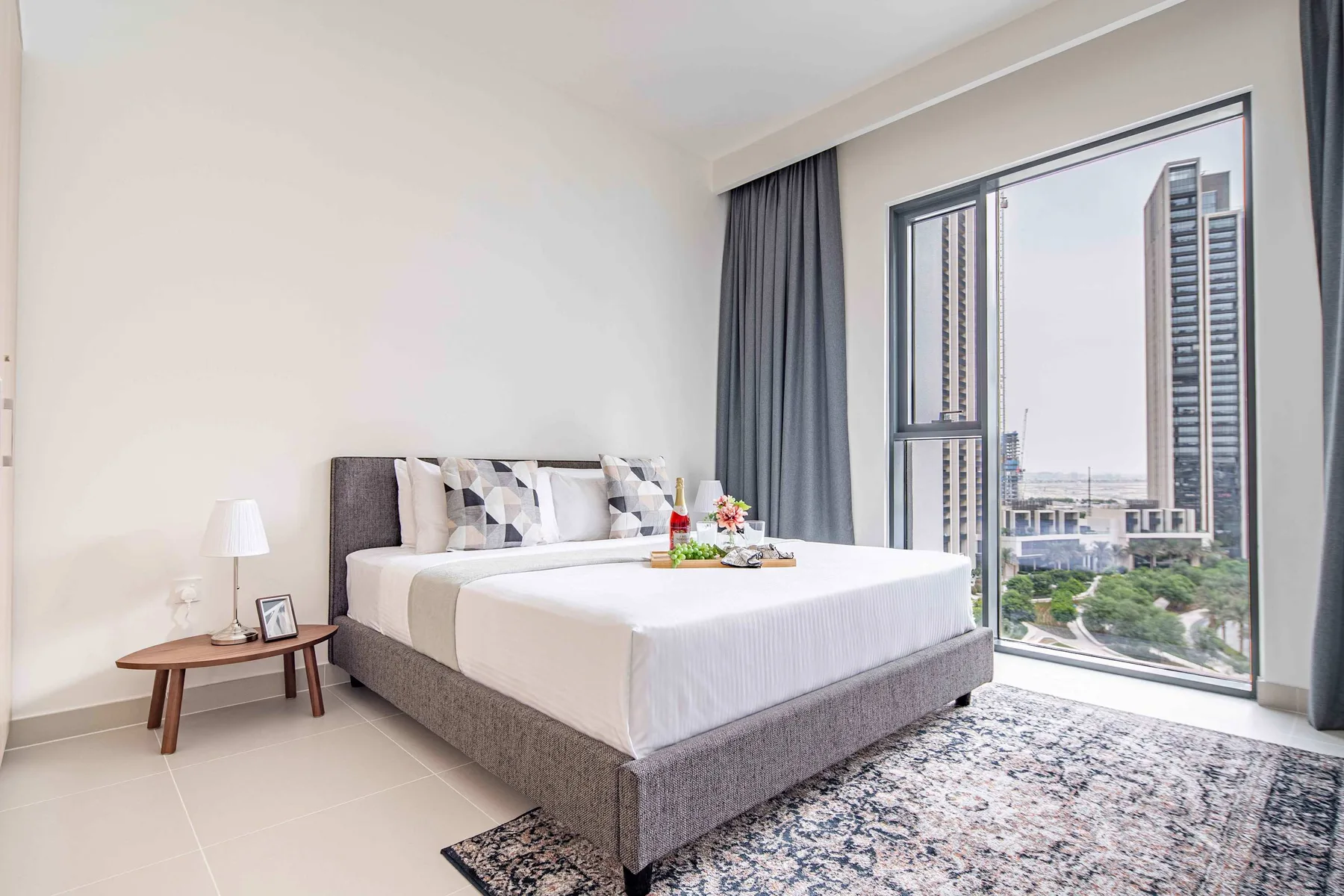 A bedroom with a bed beside a large window overlooking nearby buildings.