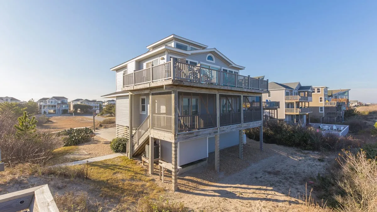 A raised coastal house with broad upper decks stands on sandy ground.