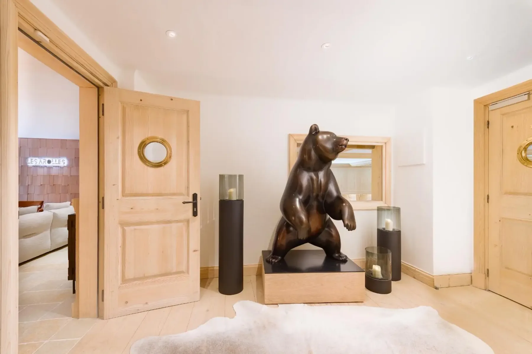 A room with a bear sculpture on a pedestal, flanked by two cylindrical candle holders. Open wooden doors lead to another room.