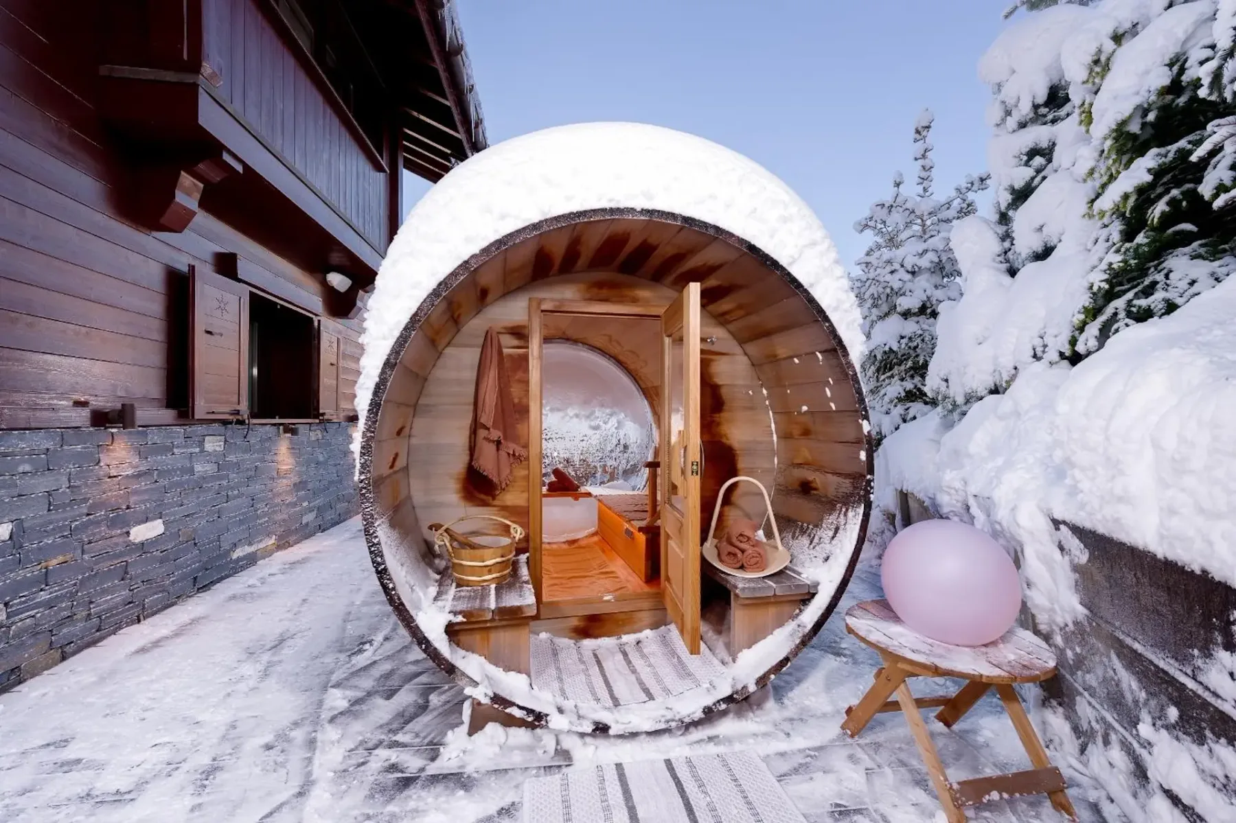 A barrel-shaped outdoor sauna covered in snow, with a cozy interior, sits on a snowy patio next to a wooden building and a small table.