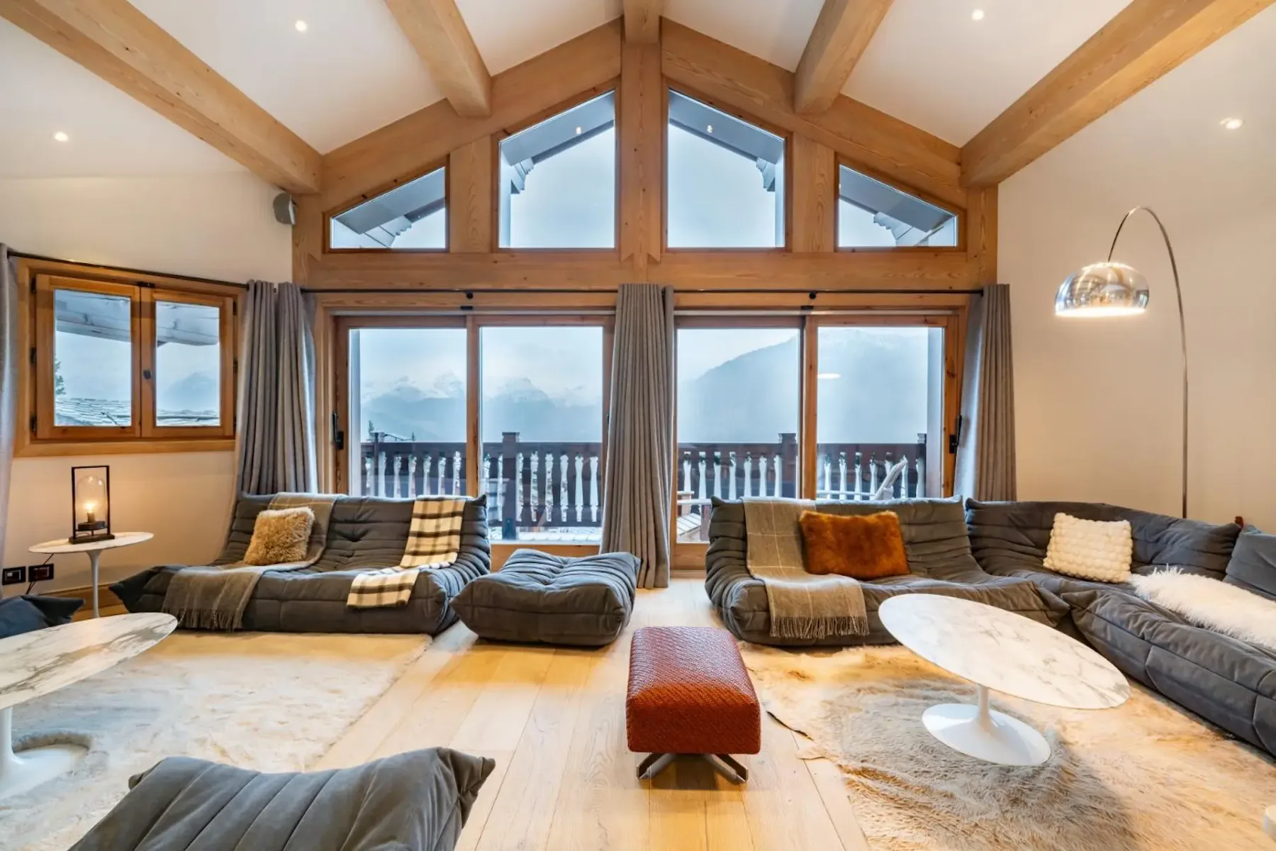 Spacious living room with large windows, mountain view, cozy sofas, a red ottoman, marble tables, and wooden beams on the ceiling.