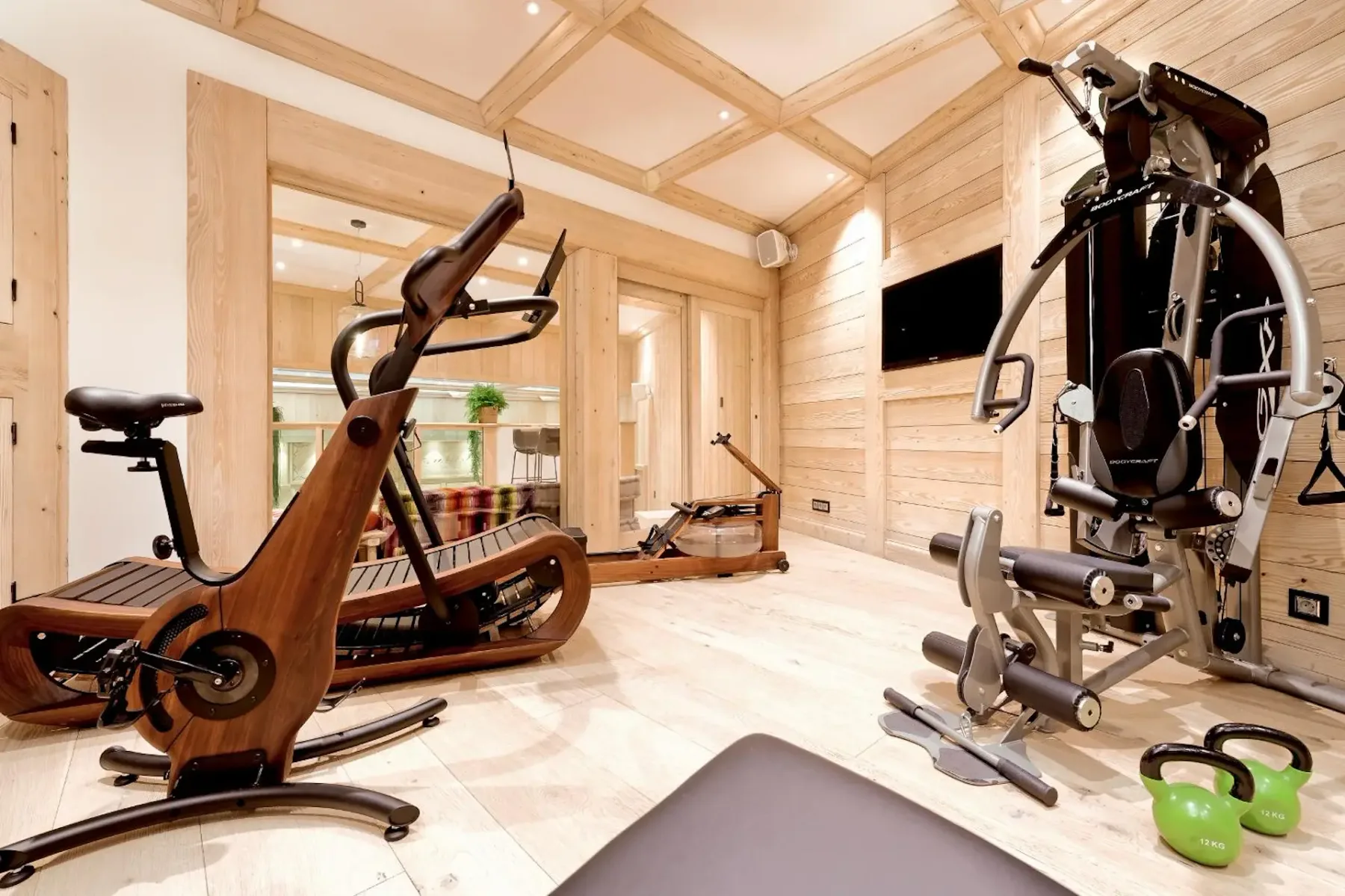 Modern home gym with a wooden exercise bike, treadmill, multi-gym machine, kettlebell, and mirrored walls on light wood flooring.