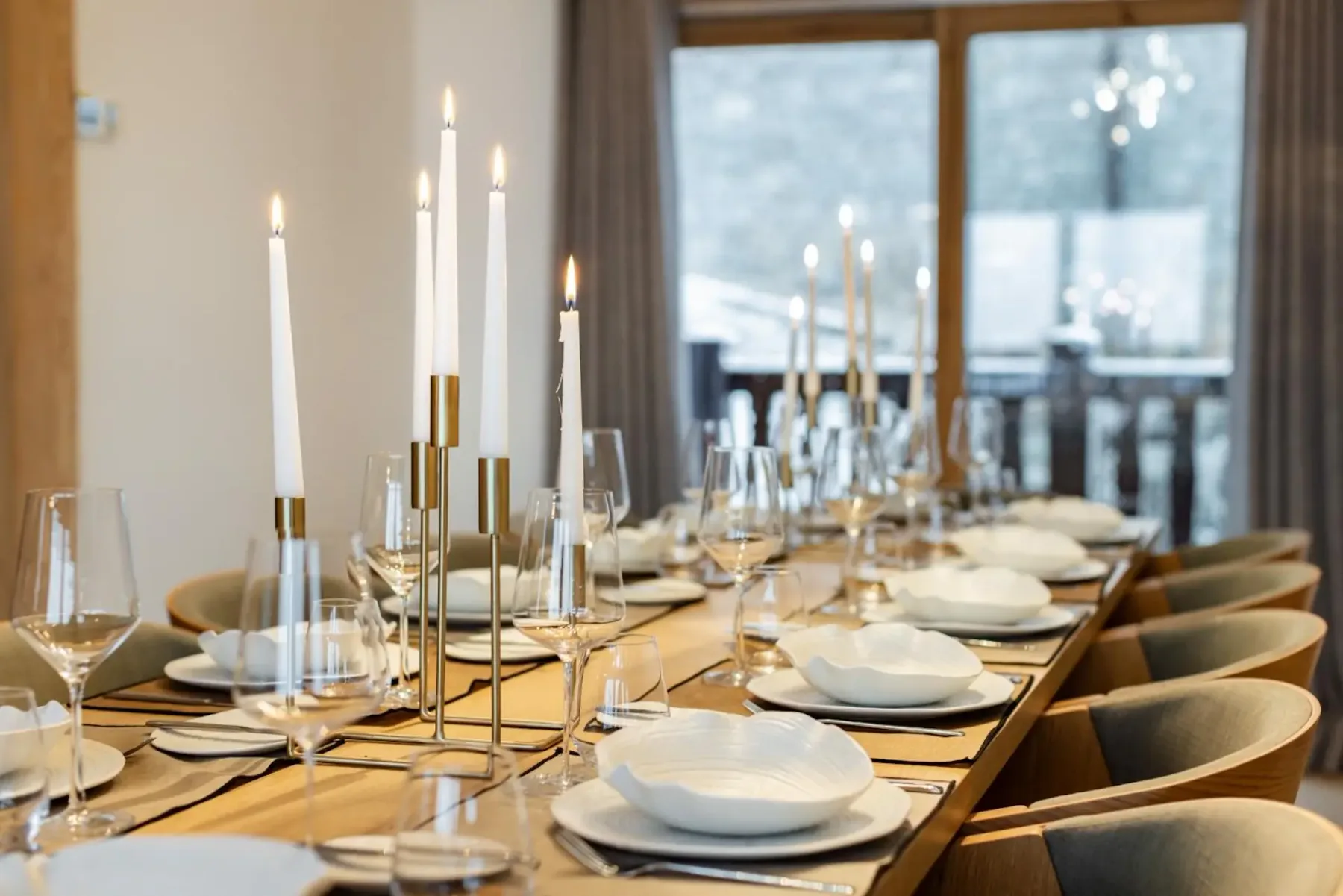 Elegant dining table set for a formal dinner, with white plates, wine glasses, and lit candlesticks, near large windows.