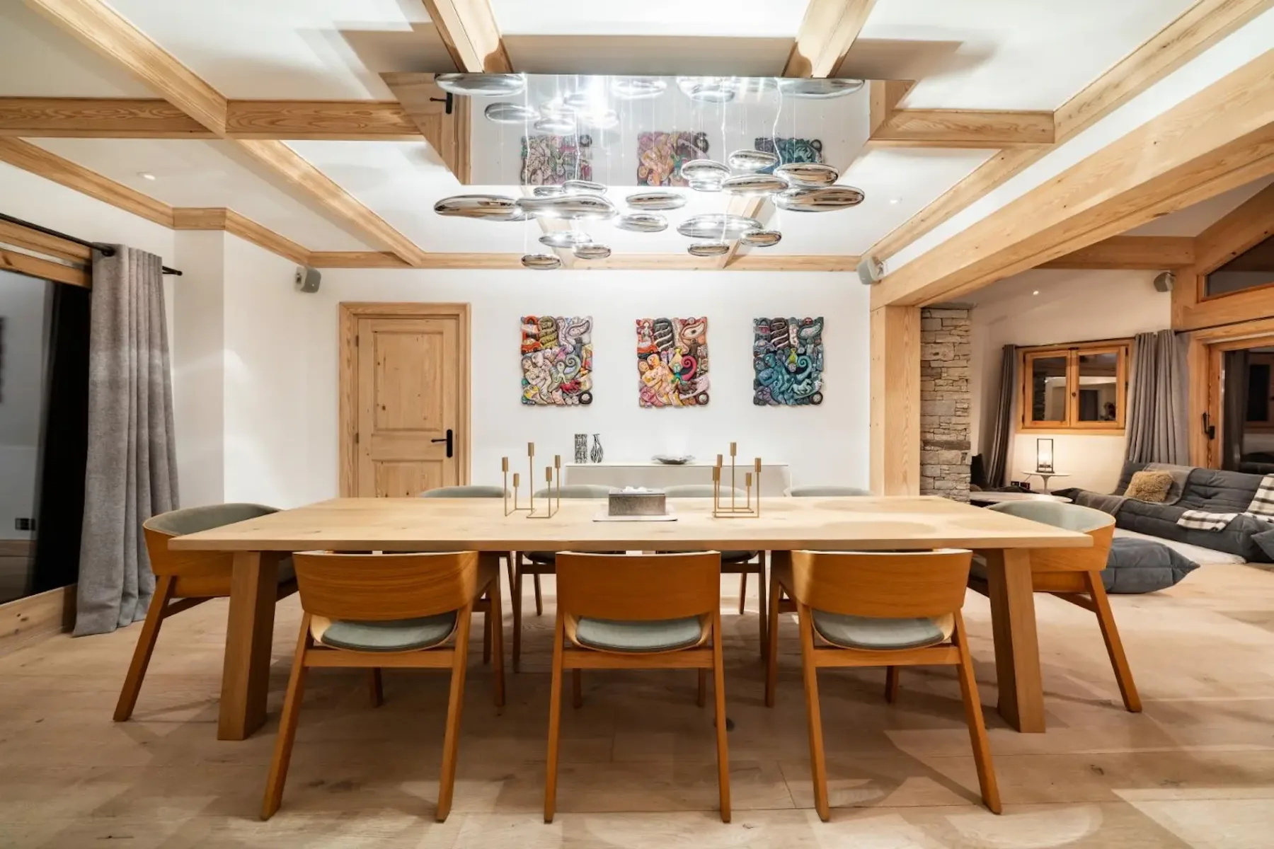 Modern dining room with a large wooden table, eight chairs, abstract wall art, and a unique overhead light fixture. Cozy seating area visible.