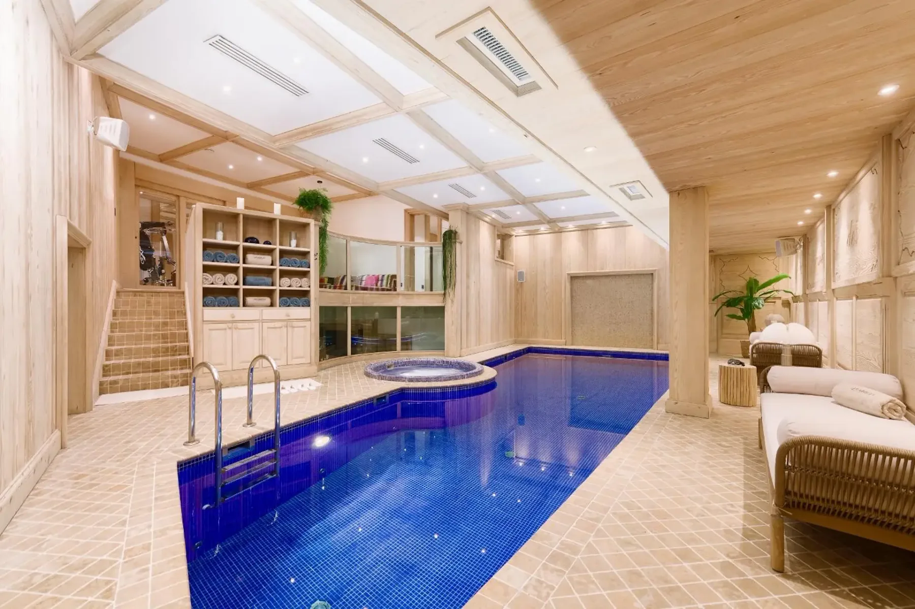 Luxurious indoor pool area with wooden walls, a blue-tiled pool, a hot tub, loungers, and shelves with towels and plants.