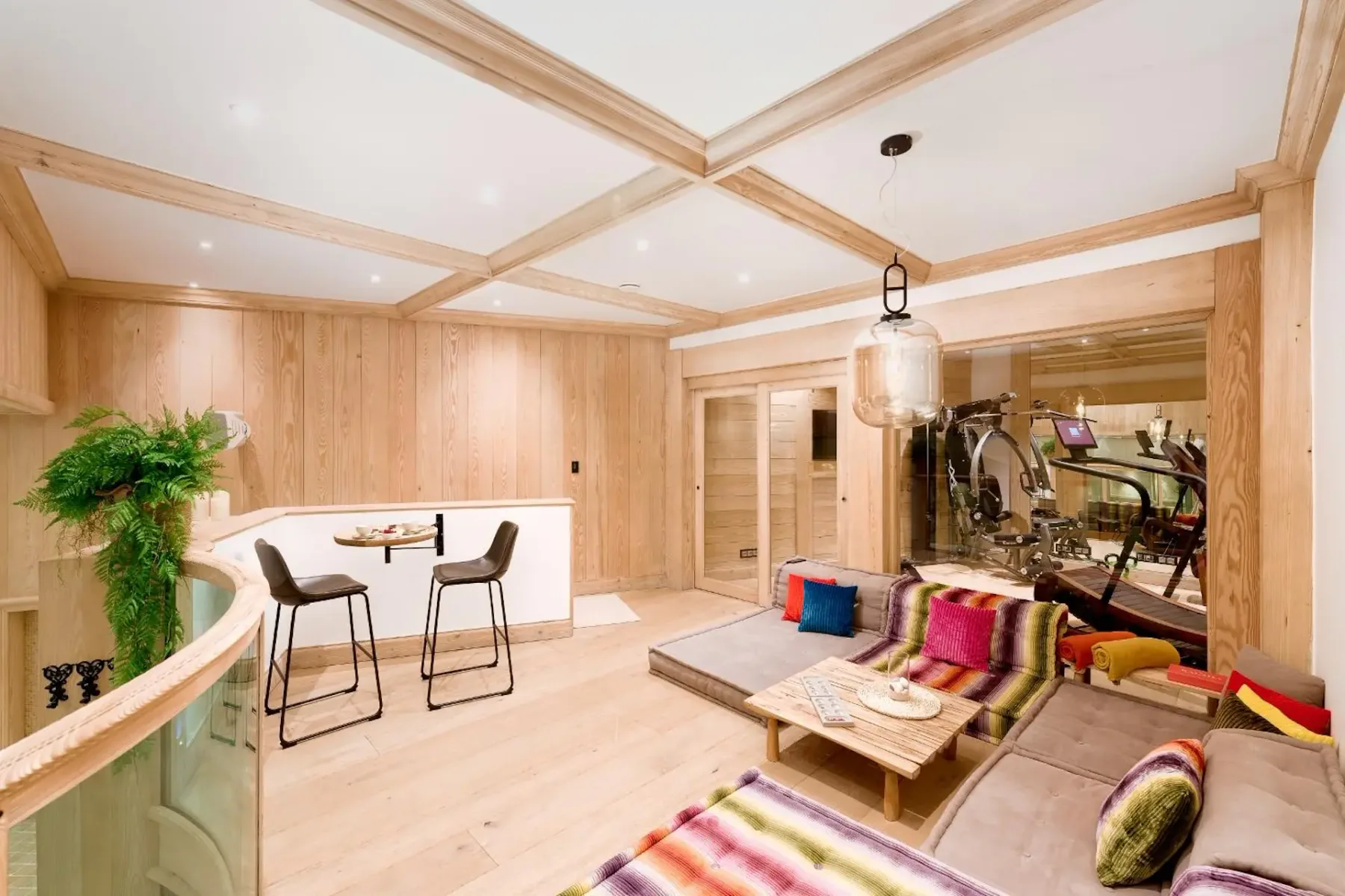 Modern living room with wooden walls, colorful cushions on a sectional sofa, a small dining table, and a view into a home gym.