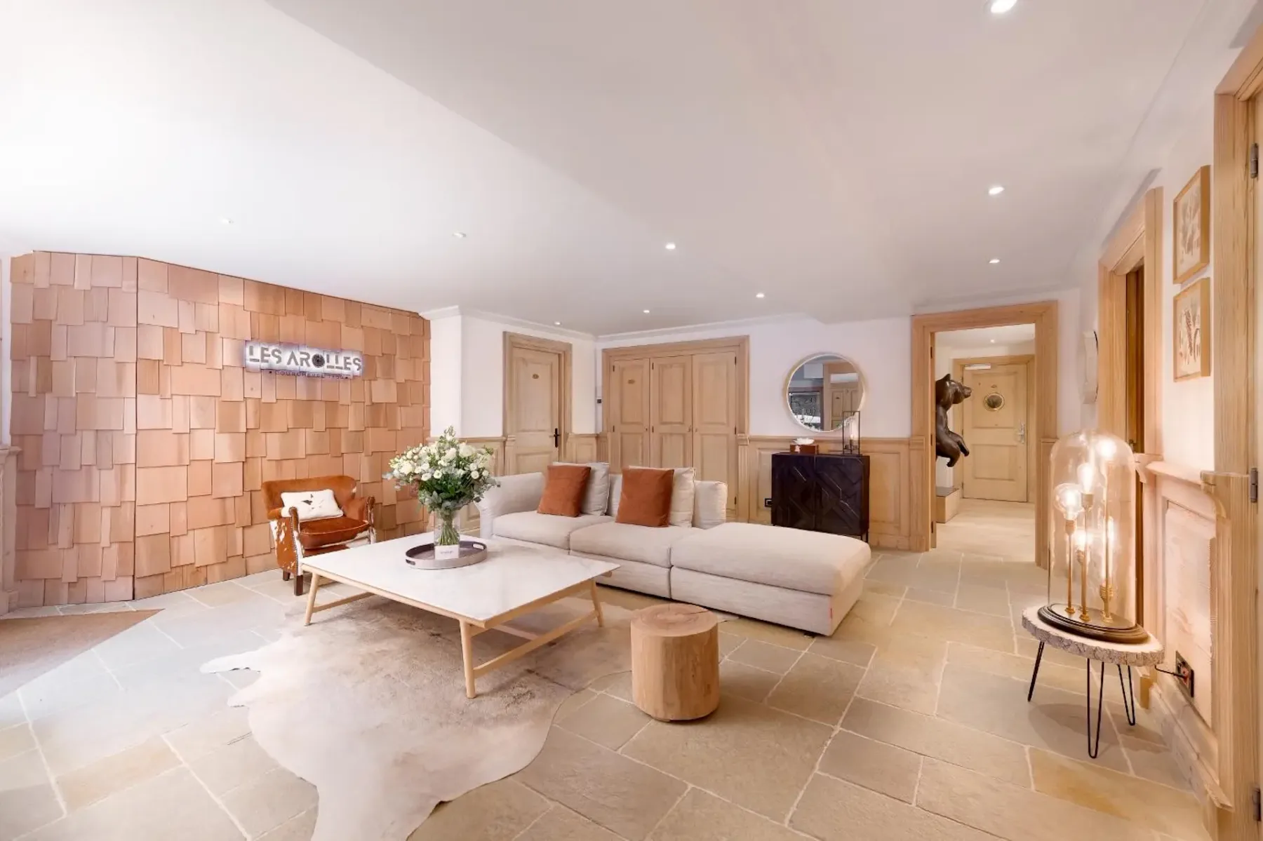 Spacious living room with a large beige sectional sofa, wooden coffee table, and decorative wall paneling. Soft lighting creates a warm ambiance.