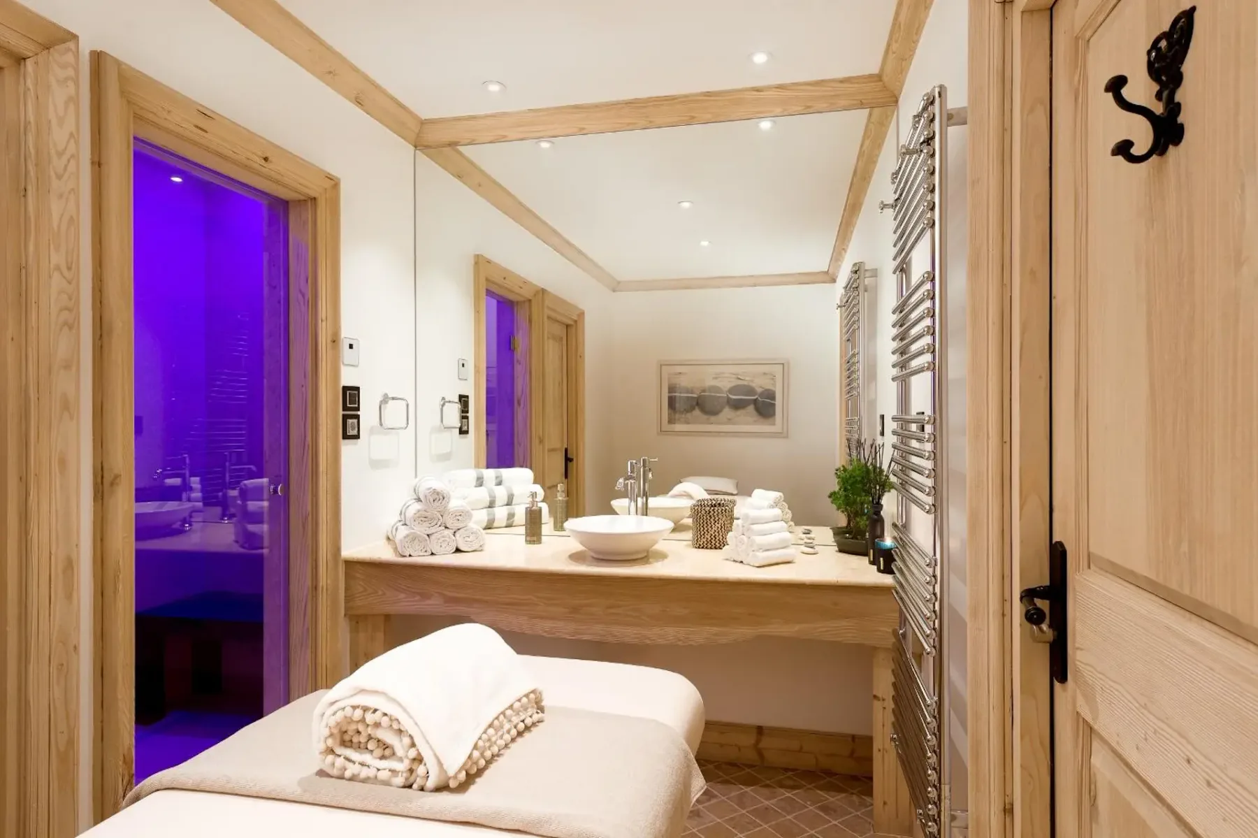 Modern spa room with a massage table, rolled towels, a sink, large mirror, and ambient purple lighting in the adjacent room.