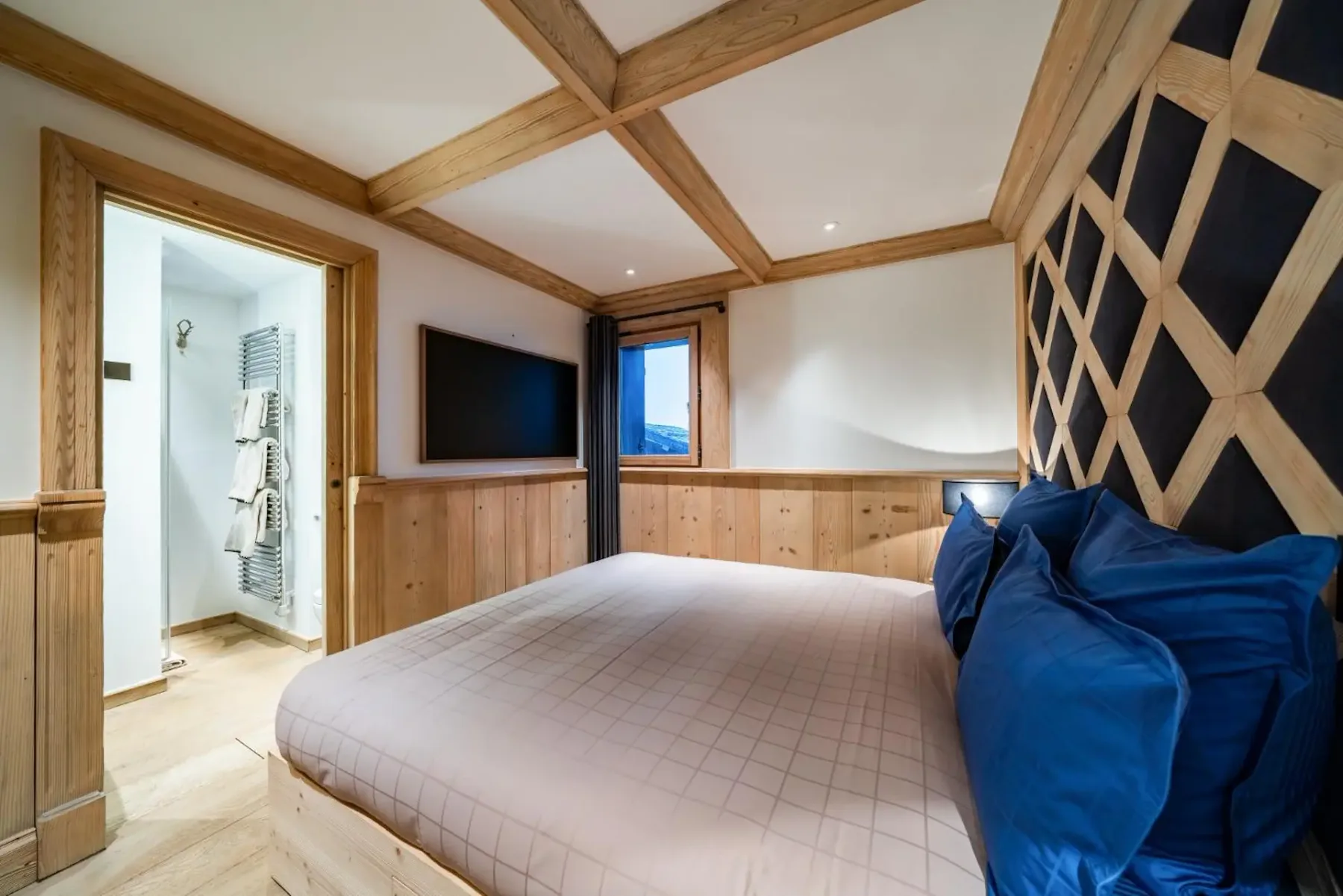 Cozy bedroom with a large bed, blue pillows, wooden accents, a mounted TV, and a view of the bathroom and window.