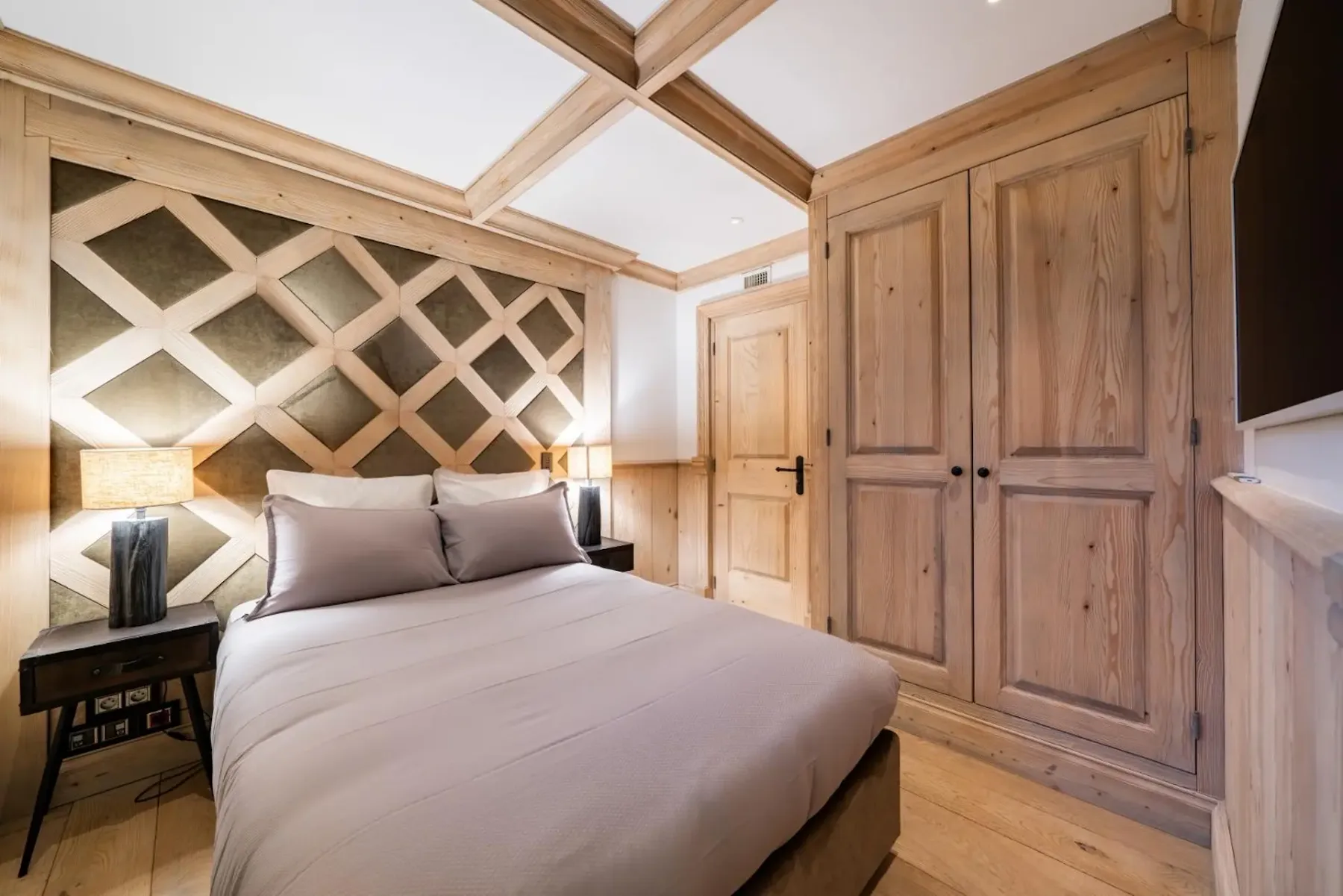 Cozy bedroom with a double bed, wooden paneling, geometric headboard, two bedside lamps, and a wooden wardrobe.