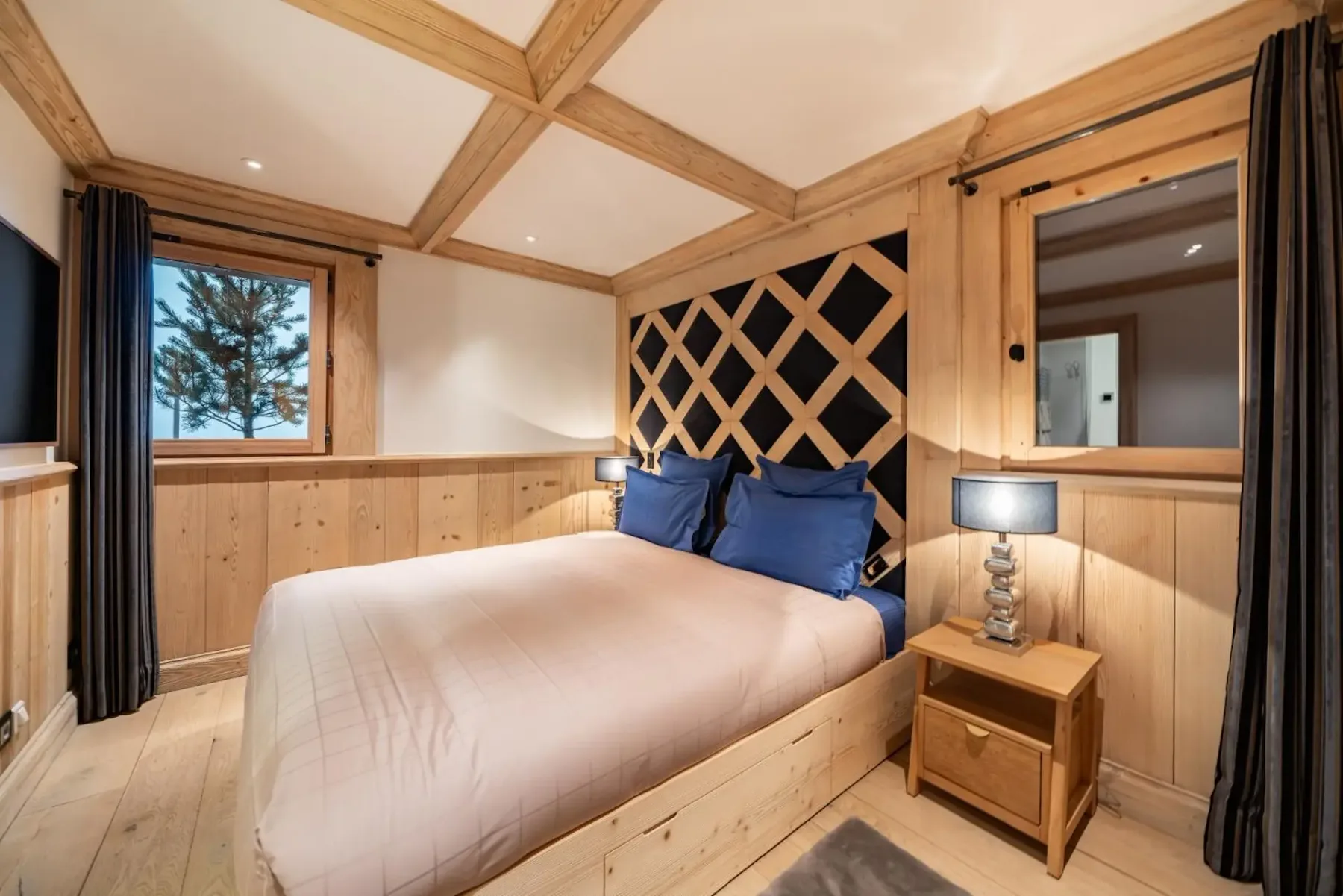 Cozy bedroom with wooden walls, a bed with blue pillows, a patterned headboard, bedside table with lamp, and a window with a tree view.