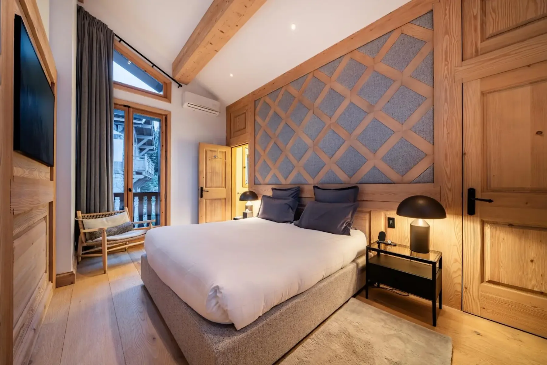 Cozy bedroom with a wooden interior, featuring a double bed, bedside tables with lamps, a chair by a window, and a textured accent wall.