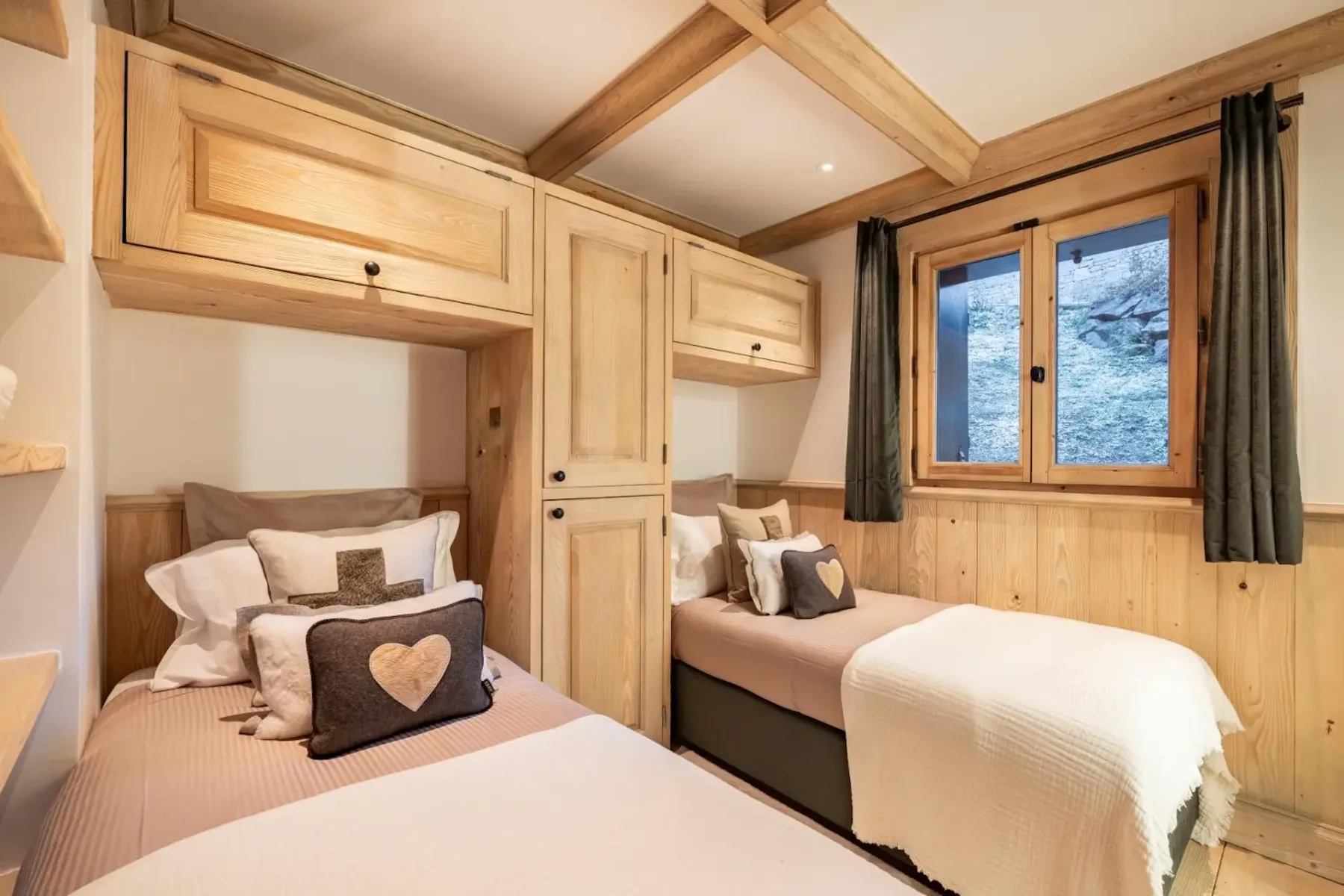 Cozy bedroom with two single beds, wooden furniture, heart-themed pillows, and a window with a view of trees outside.