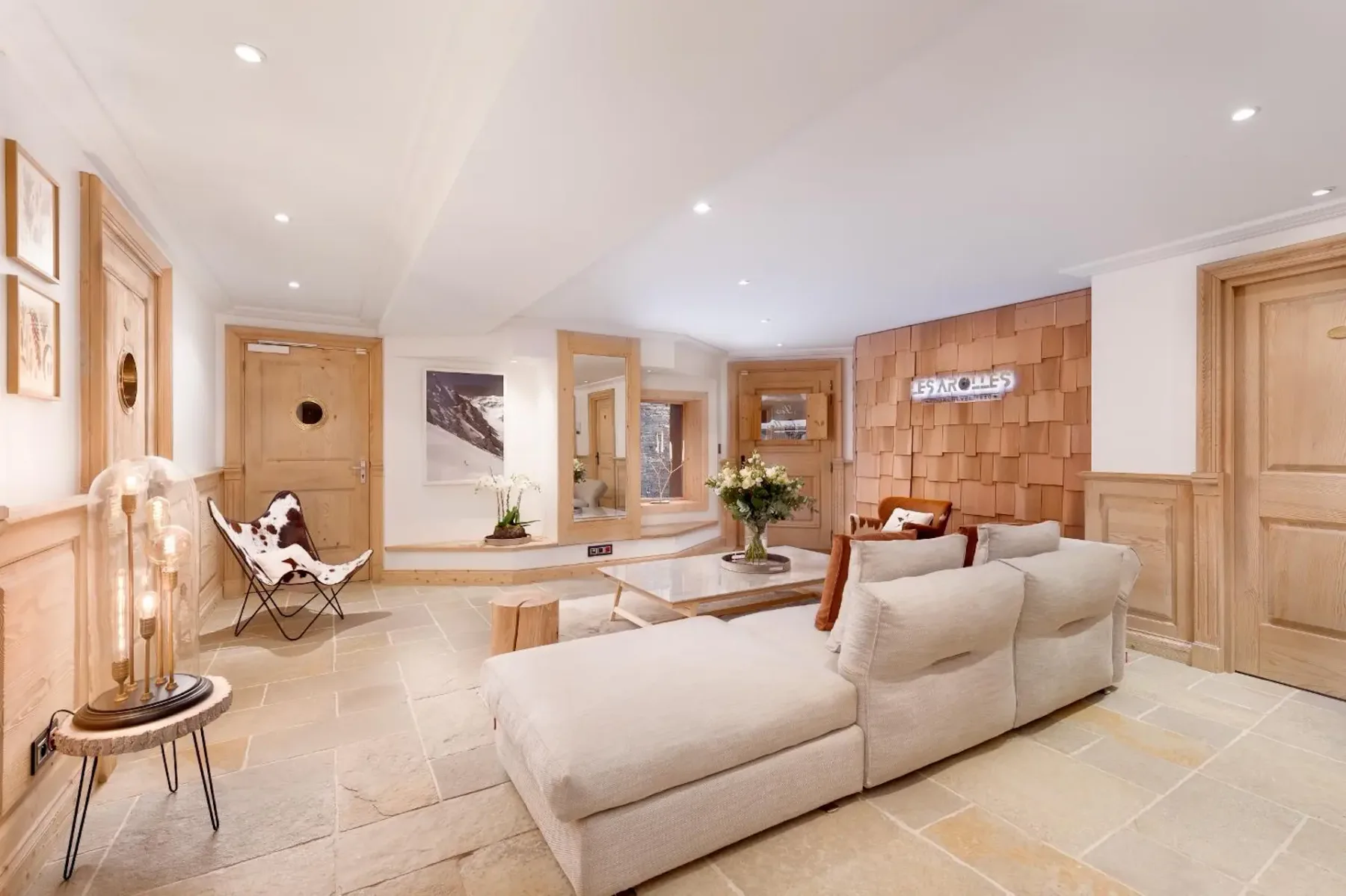 Modern living room with a cozy beige sectional sofa, wooden accents, and soft lighting. Decor includes wall art and a small table with flowers.